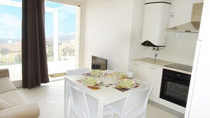 A dining area with a table set for four, a kitchen with white cabinets and a stove, and a view through a large window. - Affittimoderni Isola Rossa Élite - Pati19 (Trinità d'Agultu e Vignola)