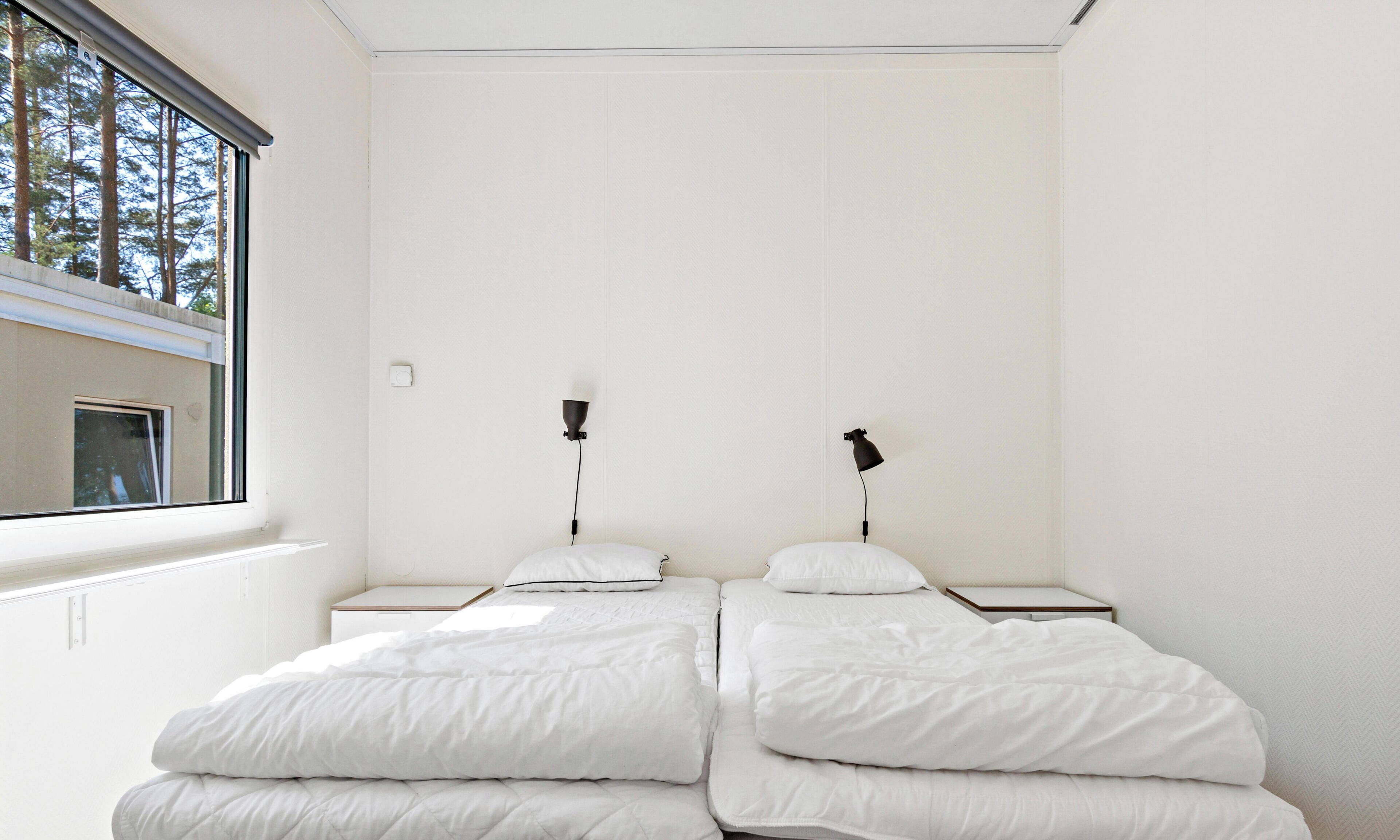 Un dormitorio con dos camas, ropa de cama blanca, una ventana y dos lámparas fijadas a la pared.