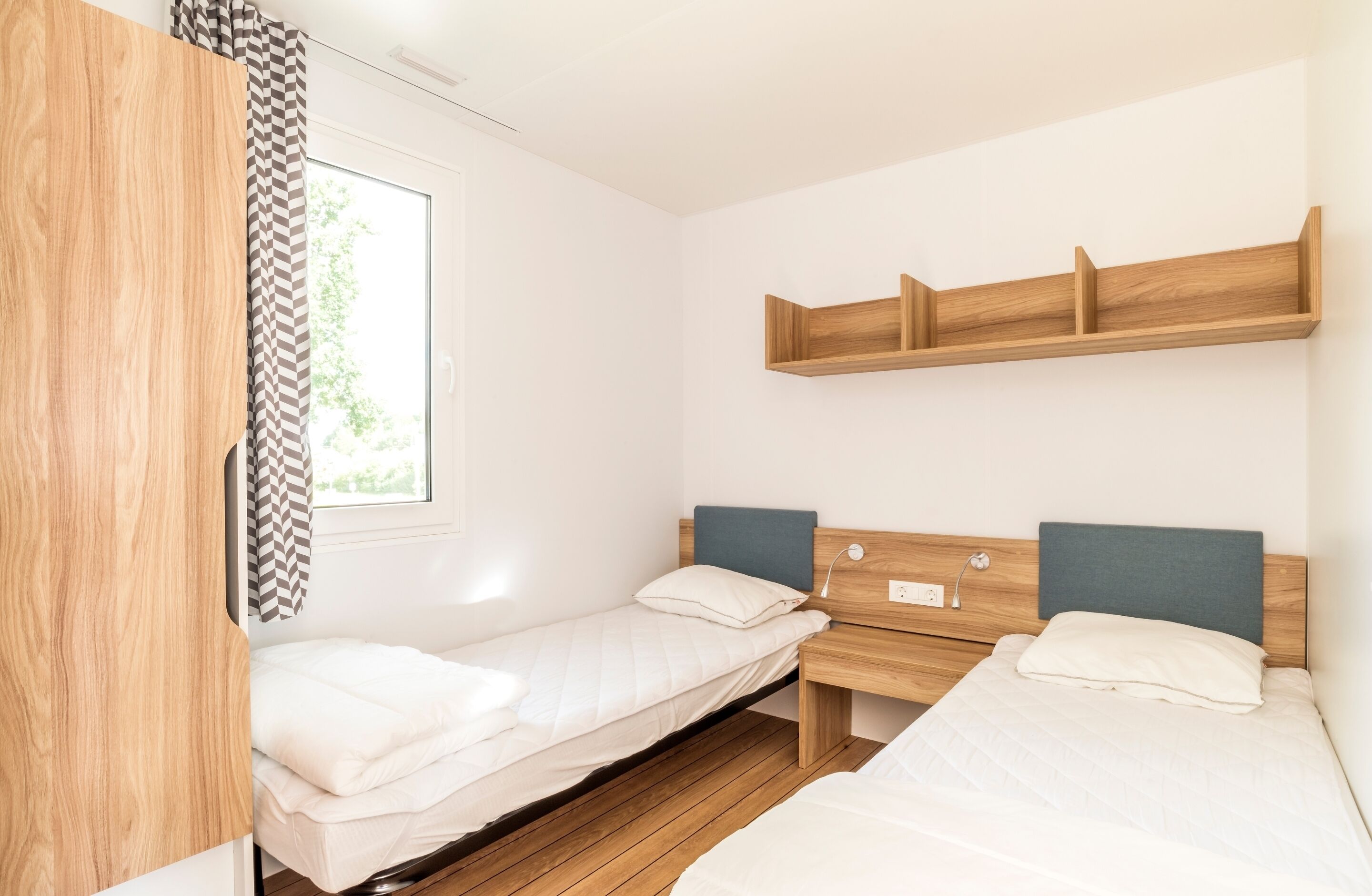 A room with two beds, a wooden wardrobe, a window with curtains, and a wall-mounted shelf.