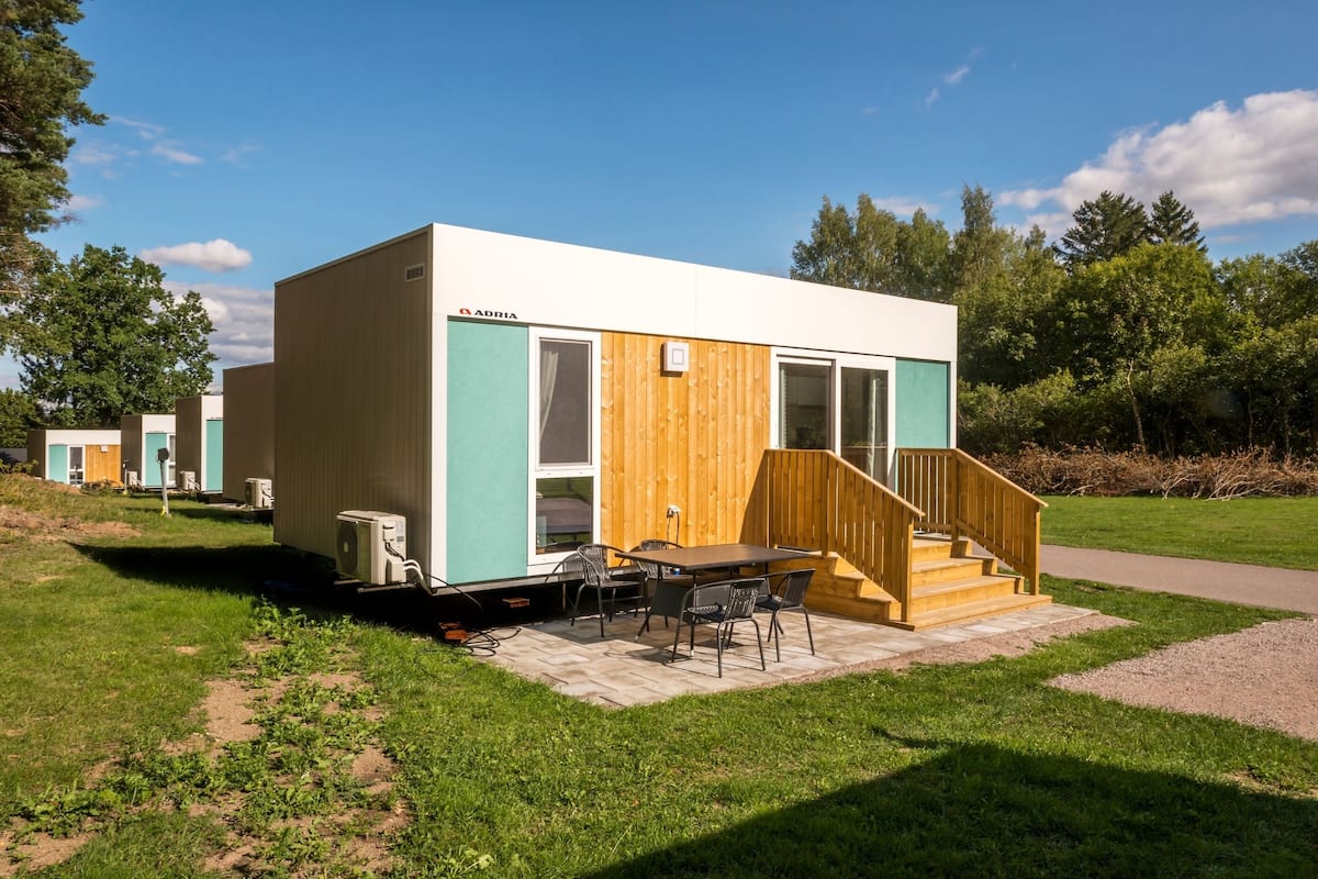 A modern, compact mobile home with a wooden staircase leading to a covered patio area.