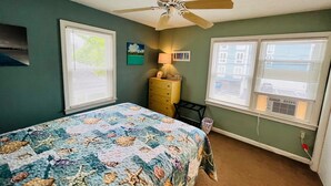 Ein Schlafzimmer mit einem Bett, einem Deckenventilator, einer Kommode und zwei Fenstern mit Jalousien.