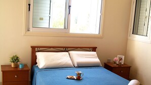 A neatly made bed with a blue bedspread, two pillows, and two rolled towels on the foot of the bed. There is a small table with a plant and a cup next to the bed. - Villa Nektaria Green Bay DPSV308 (Paralimni)