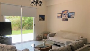 A living room with a sofa, a patterned armchair, a glass coffee table, and a television. - Villa Nektaria Green Bay DPSV308 (Paralimni)