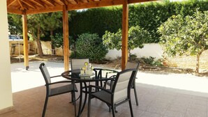 A covered outdoor patio with a round table and four chairs, surrounded by a garden with various plants and trees. - Villa Nektaria Green Bay DPSV308 (Paralimni)