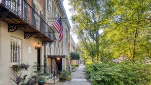 A narrow urban street with a building featuring a balcony and an American flag. - The Sarah Elizabeth House on beautiful Jones St w/ Private Courtyard (Savannah)