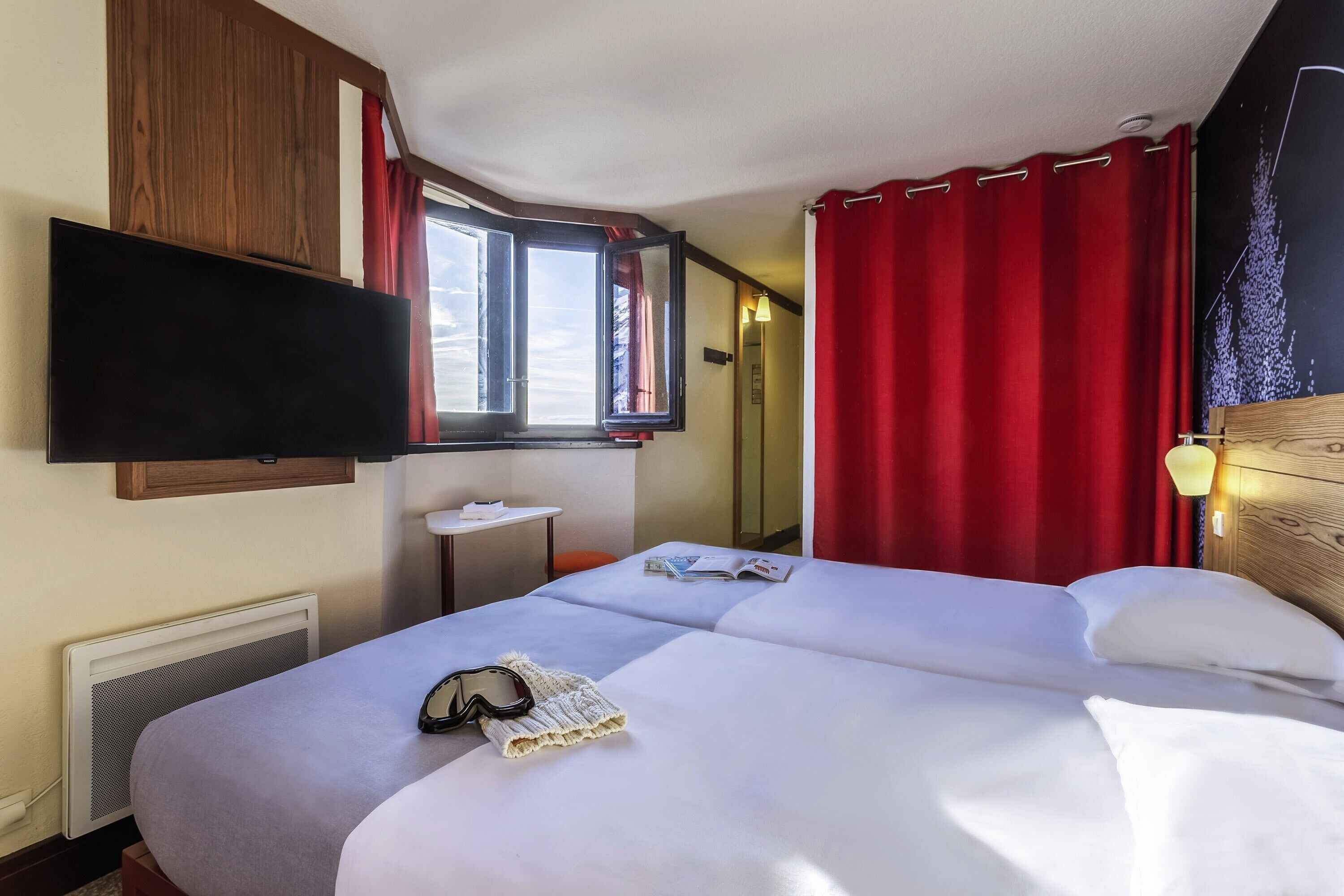 A hotel room with a bed, a TV mounted on the wall, a small table, and a window with red curtains.