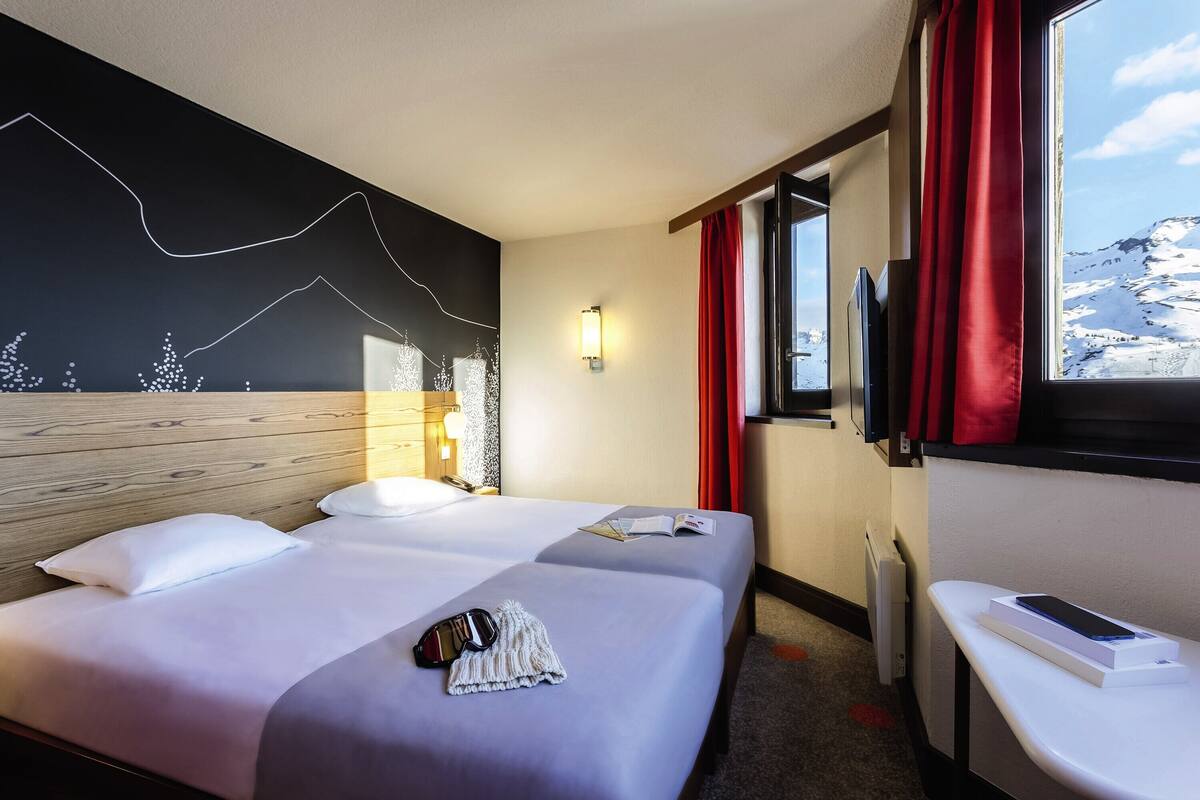 A hotel room with two beds, a TV, a window with red curtains, and a view of snow-capped mountains.