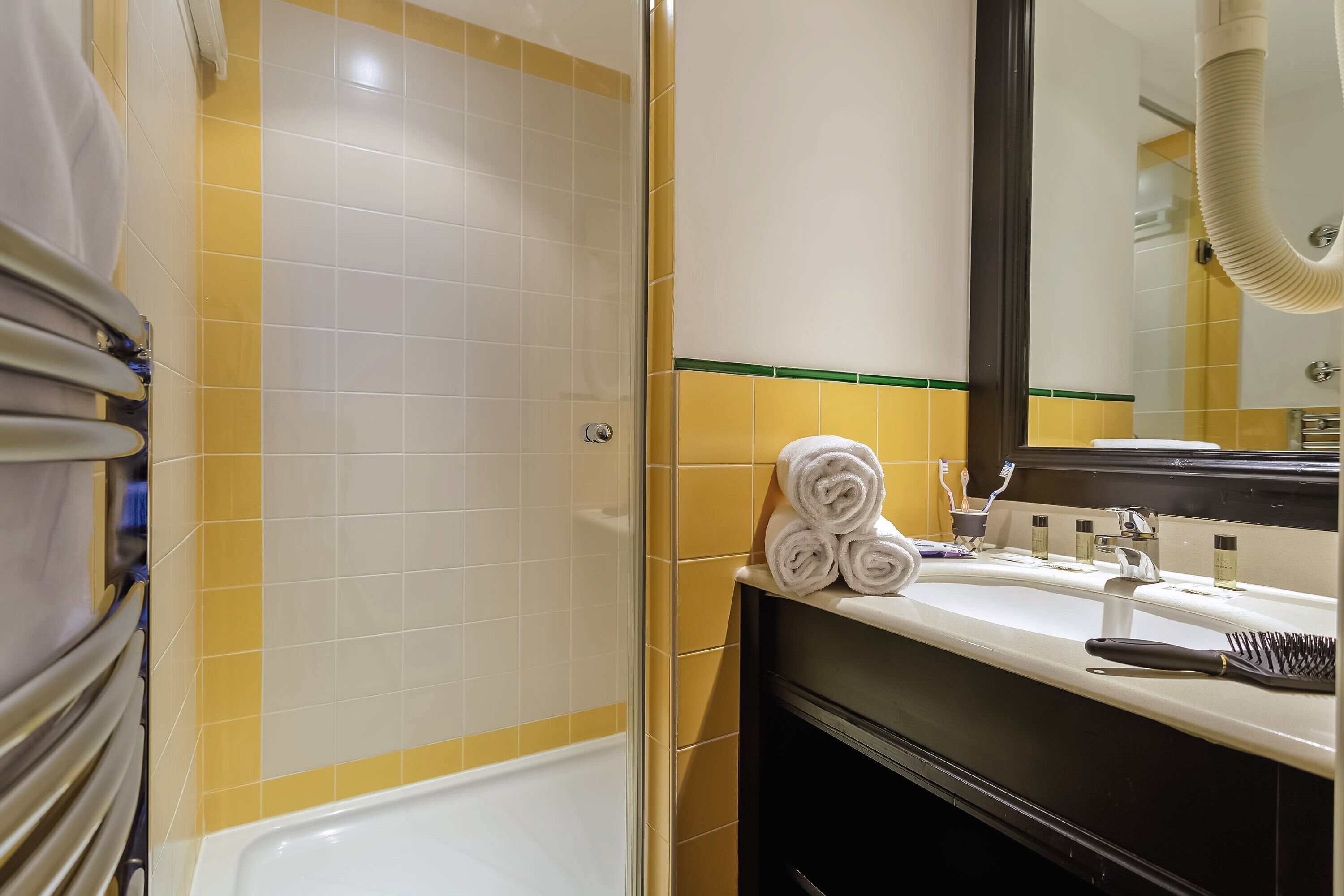 Une salle de bain avec une baignoire blanche, des carreaux jaunes et blancs, un lavabo avec un miroir et un porte-serviettes avec des serviettes pliées.
