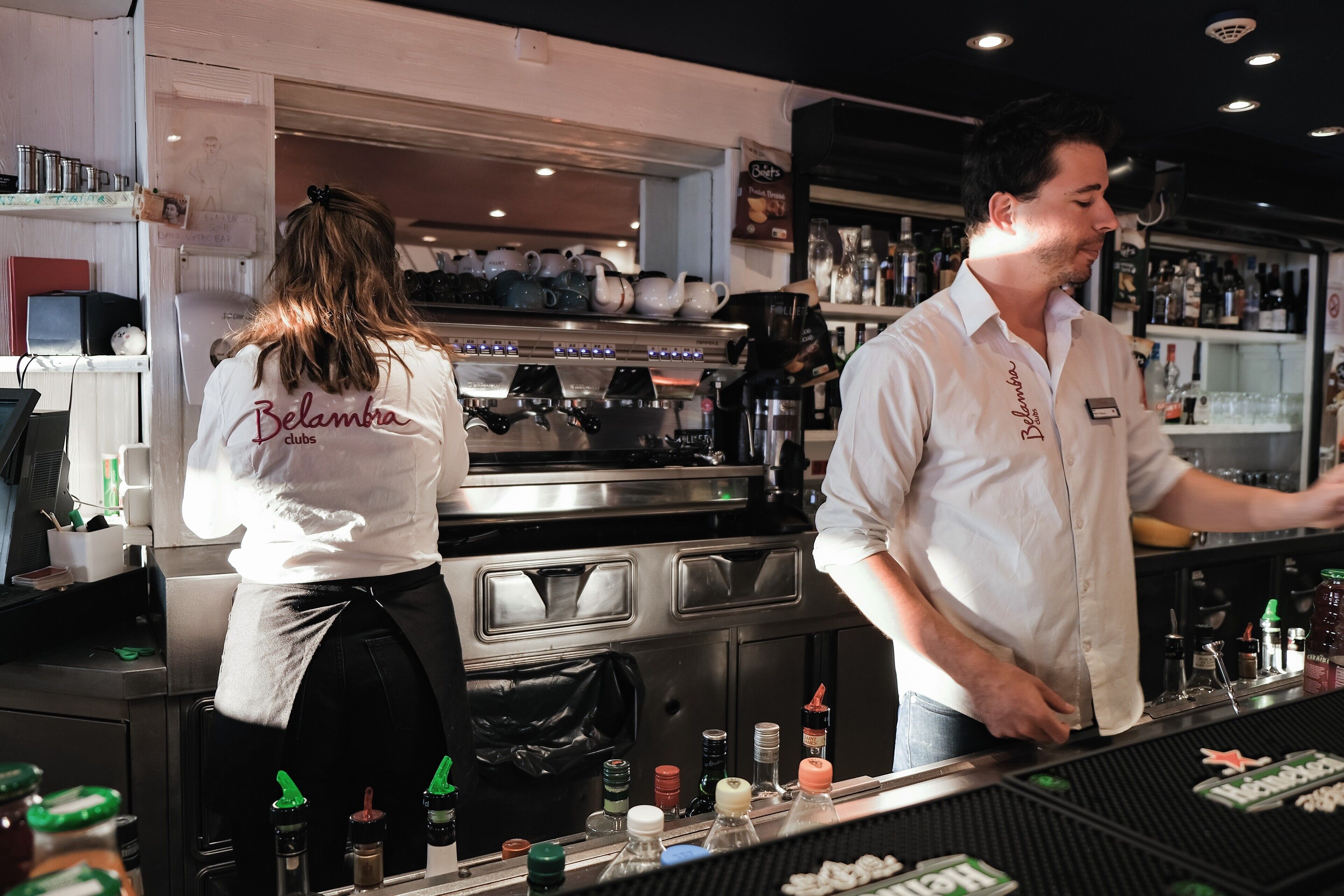 A bar area with a coffee machine, shelves with bottles, and a counter with a name tag.