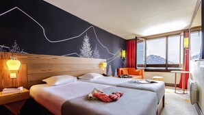 A hotel room with two beds, a wooden headboard, a wall mural, a window with a mountain view, and a small table with a chair. - Belambra Clubs Avoriaz - Les Cimes du Soleil  (Morzine)