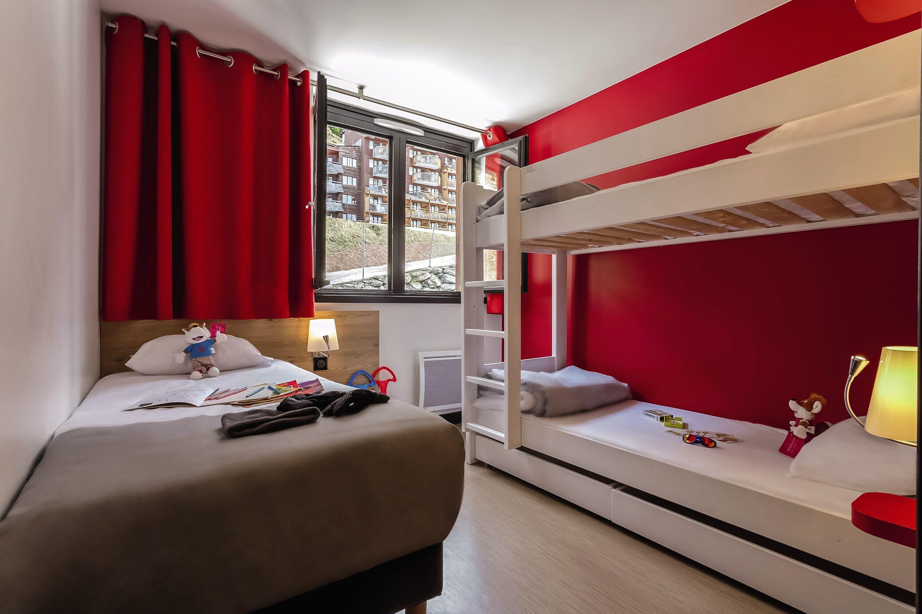 A room with a bunk bed, a desk with a lamp, and a window with red curtains.