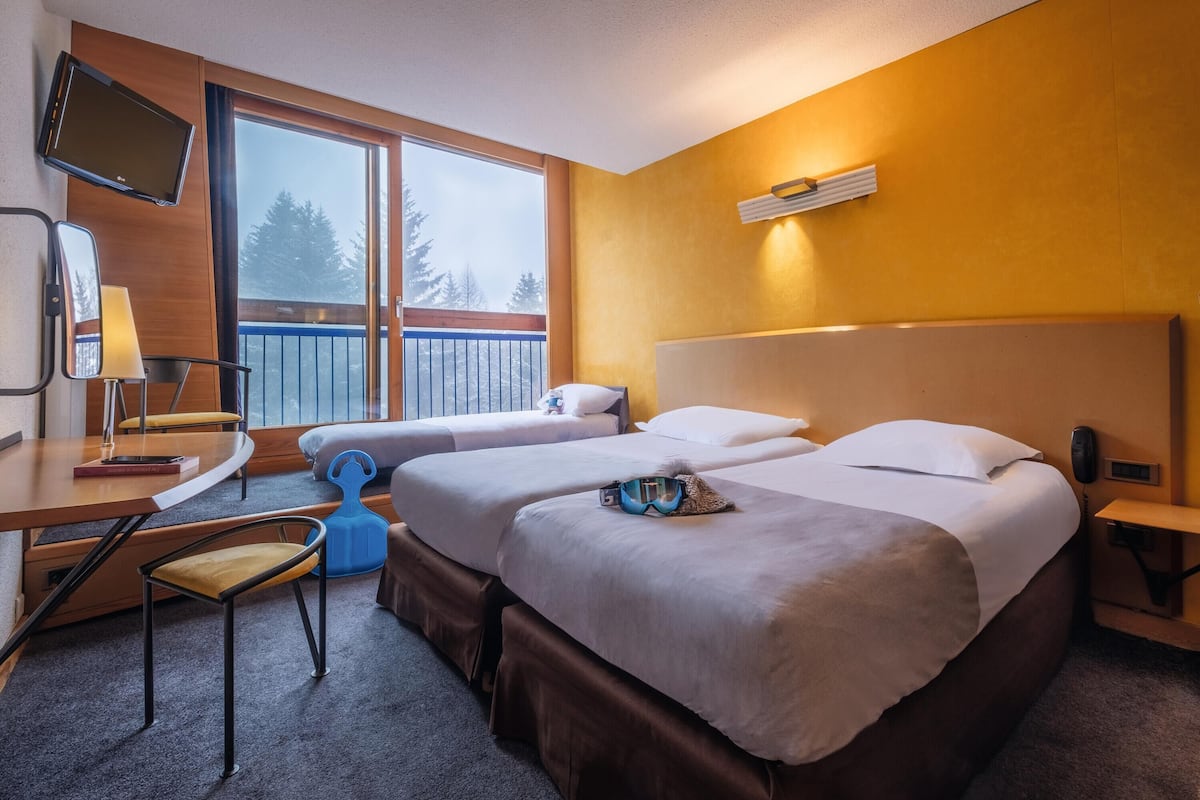 A hotel room with two beds, a desk, a chair, a TV, and a balcony with a view of trees.