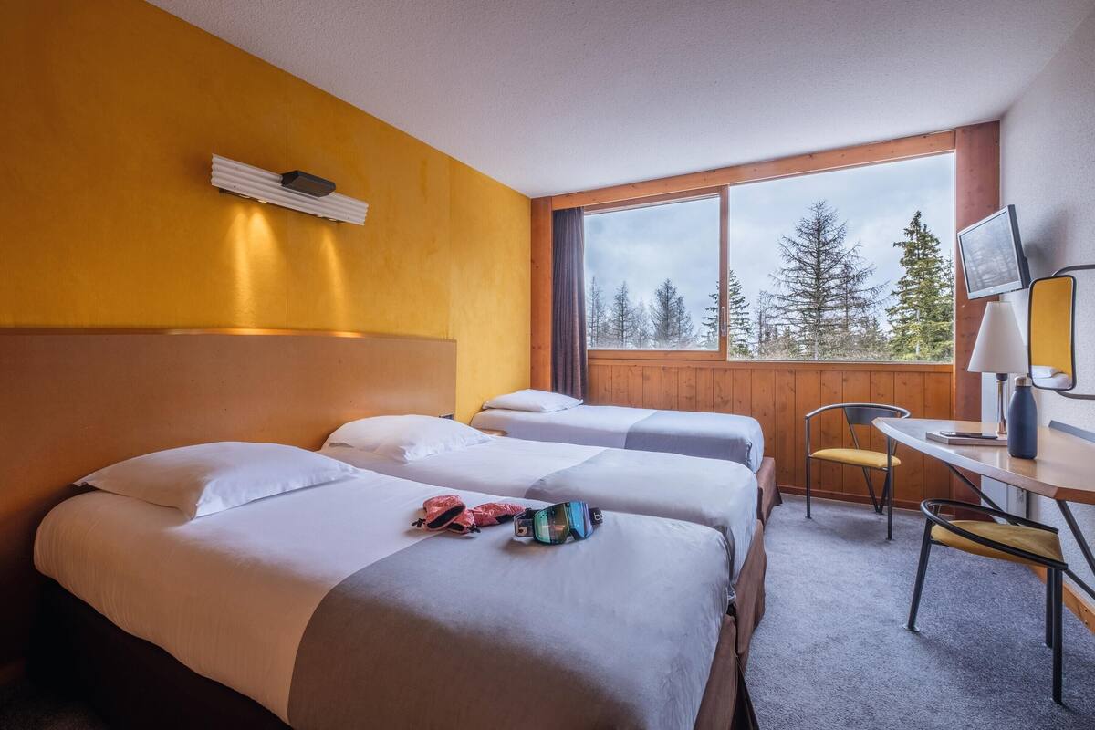 A hotel room with two beds, a desk, a chair, and a window with a view of trees.