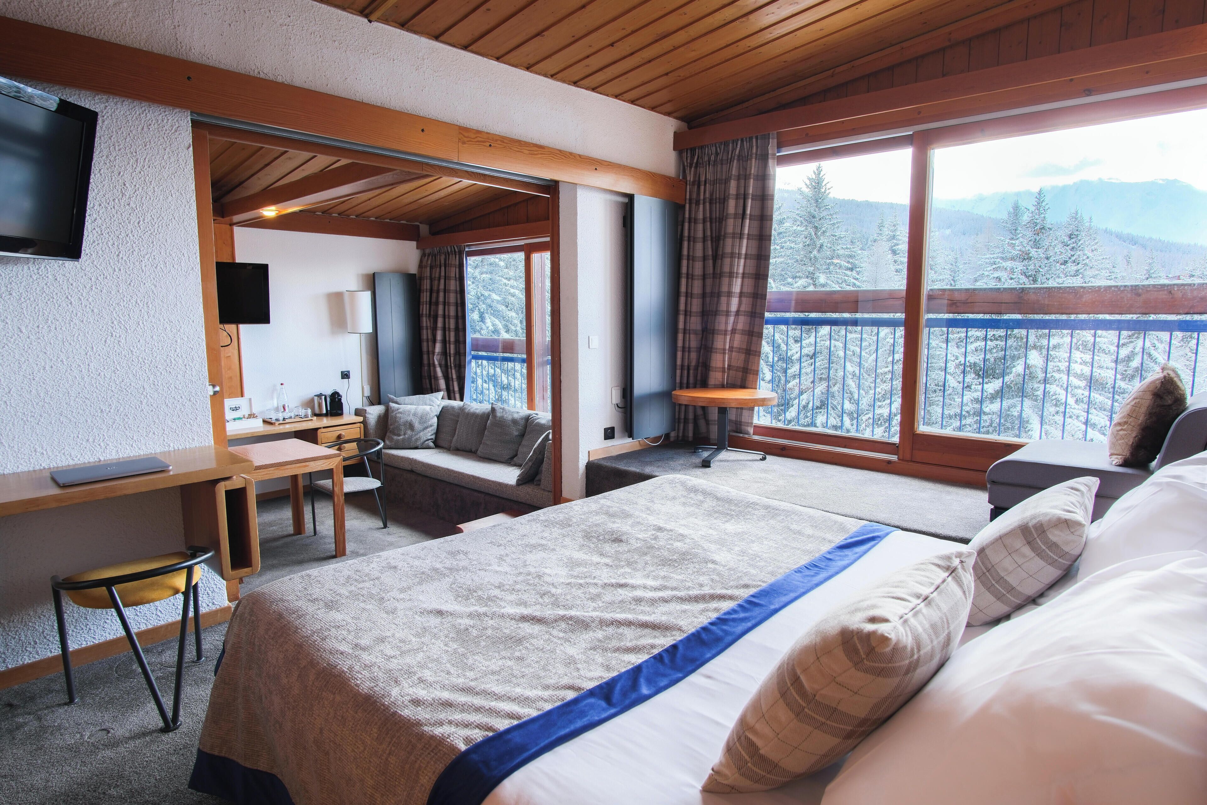 Habitación de hotel con una cama grande, un escritorio, una silla y un televisor. Ventanas con vistas a árboles nevados y montañas.