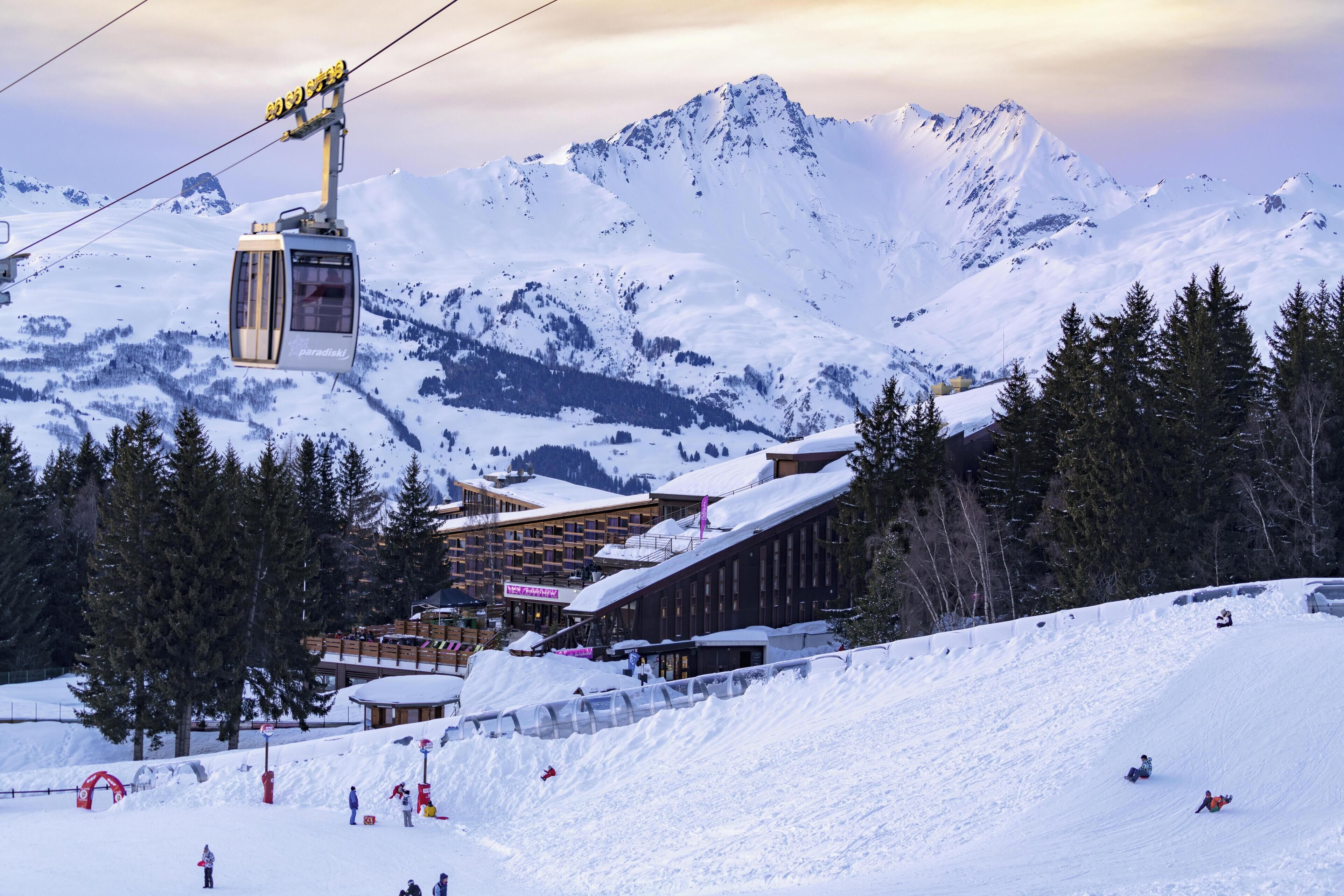 A ski lift with a cable car moving over a snowy mountain. There are buildings with snow-covered roofs and a ski slope with skiers.