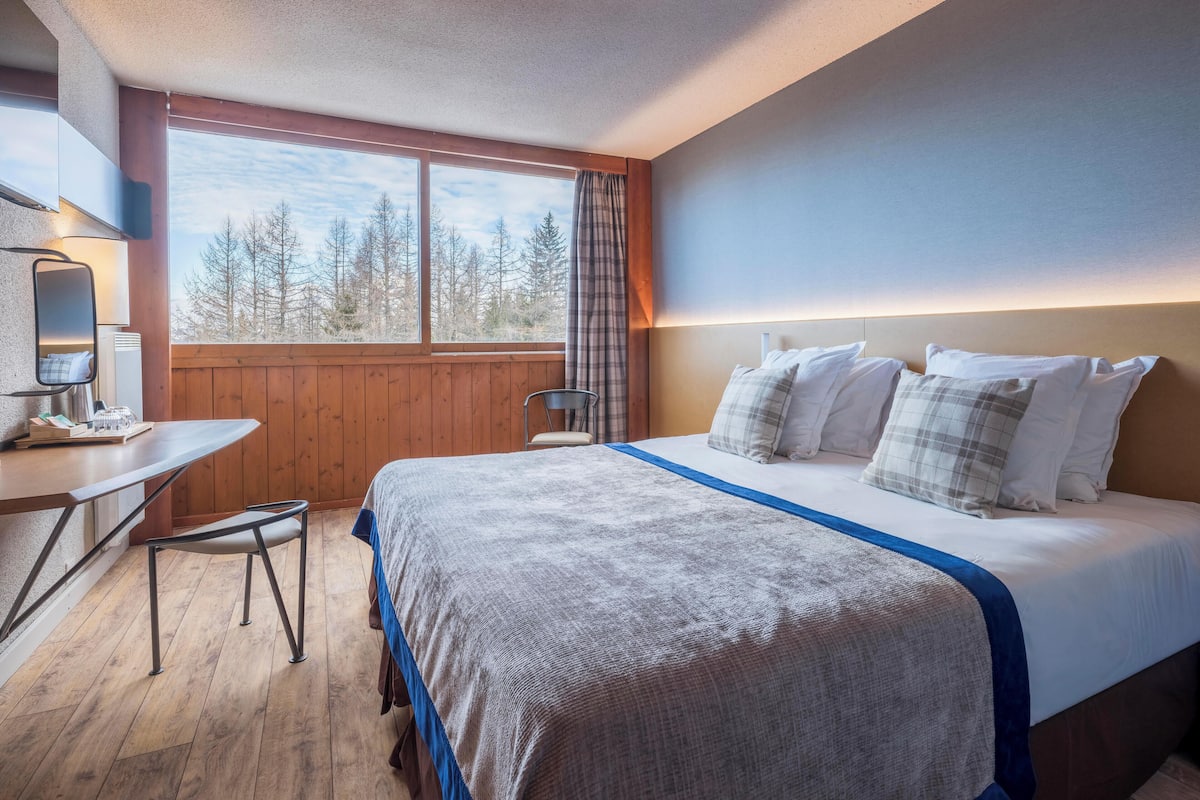 A bedroom with a large bed, a desk, and a window with a view of trees.