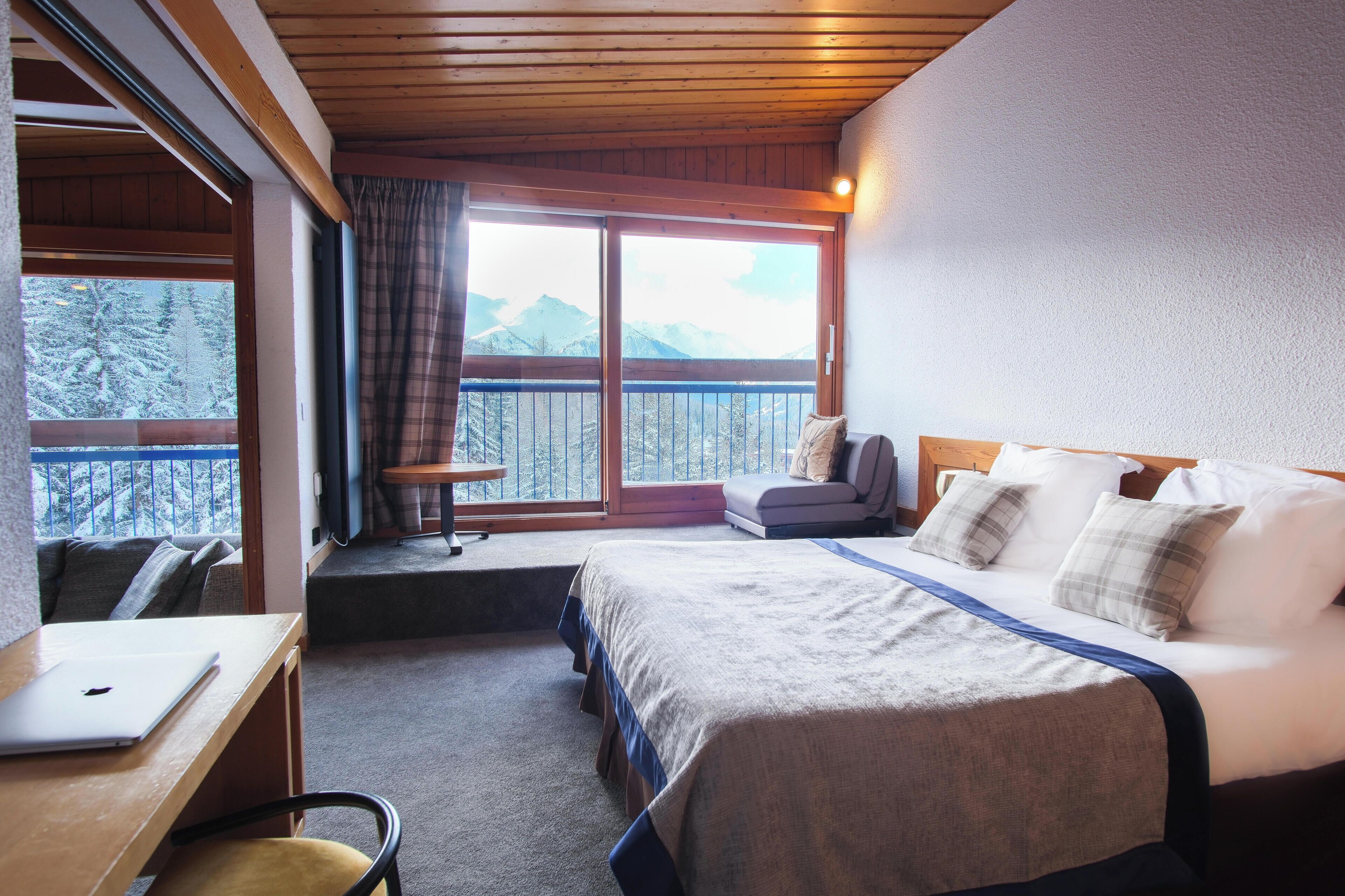 A bedroom with a large bed, a desk with a laptop, a chair, and a view of snow-covered mountains through a balcony door.