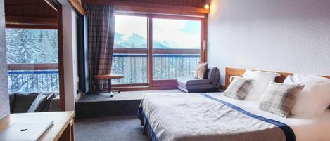 A bedroom with a large bed, a desk with a laptop, a chair, and a view of snow-covered mountains through a balcony door.