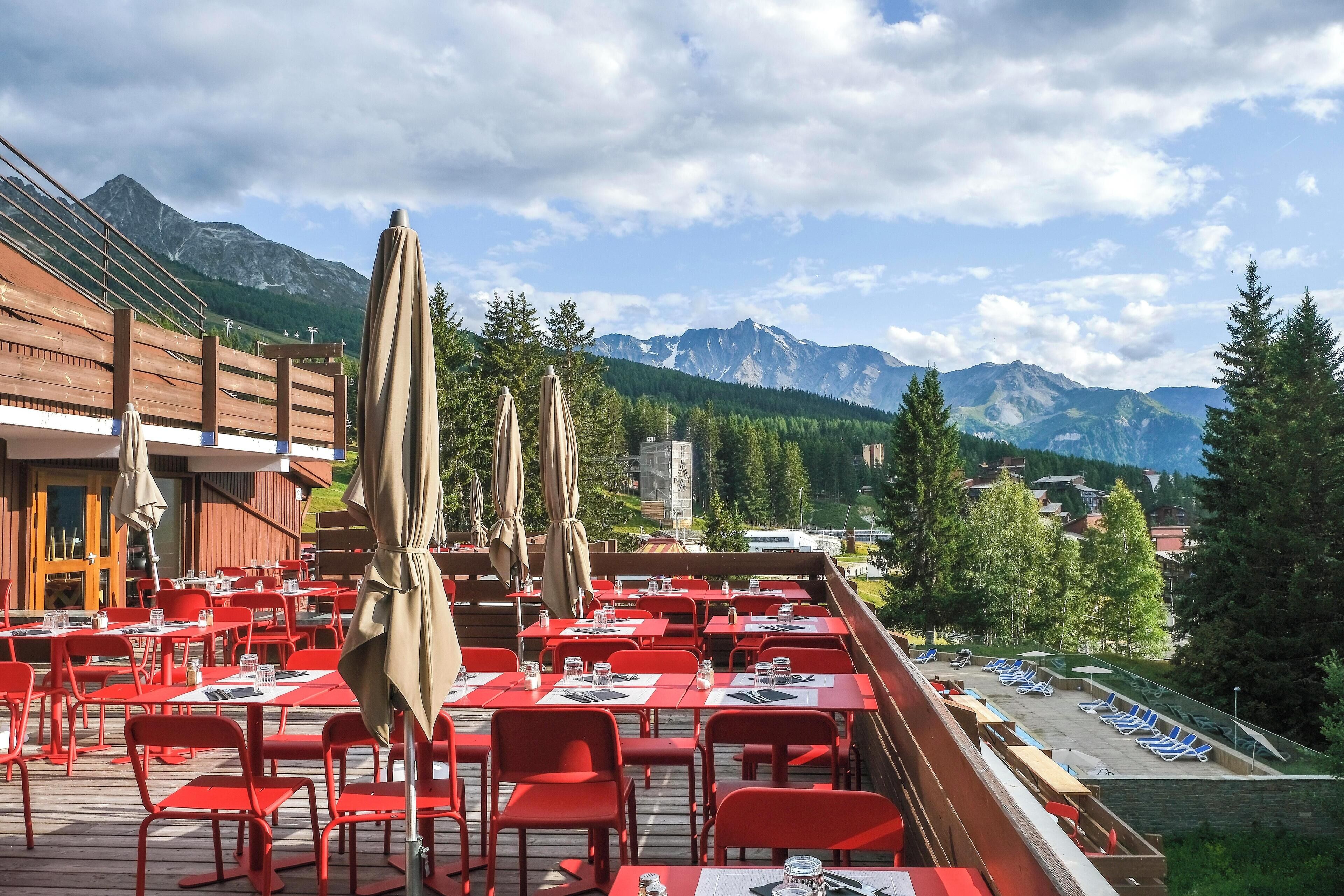 Un espace de restauration en plein air avec des tables et des chaises rouges, des parasols beiges et une vue sur la montagne.