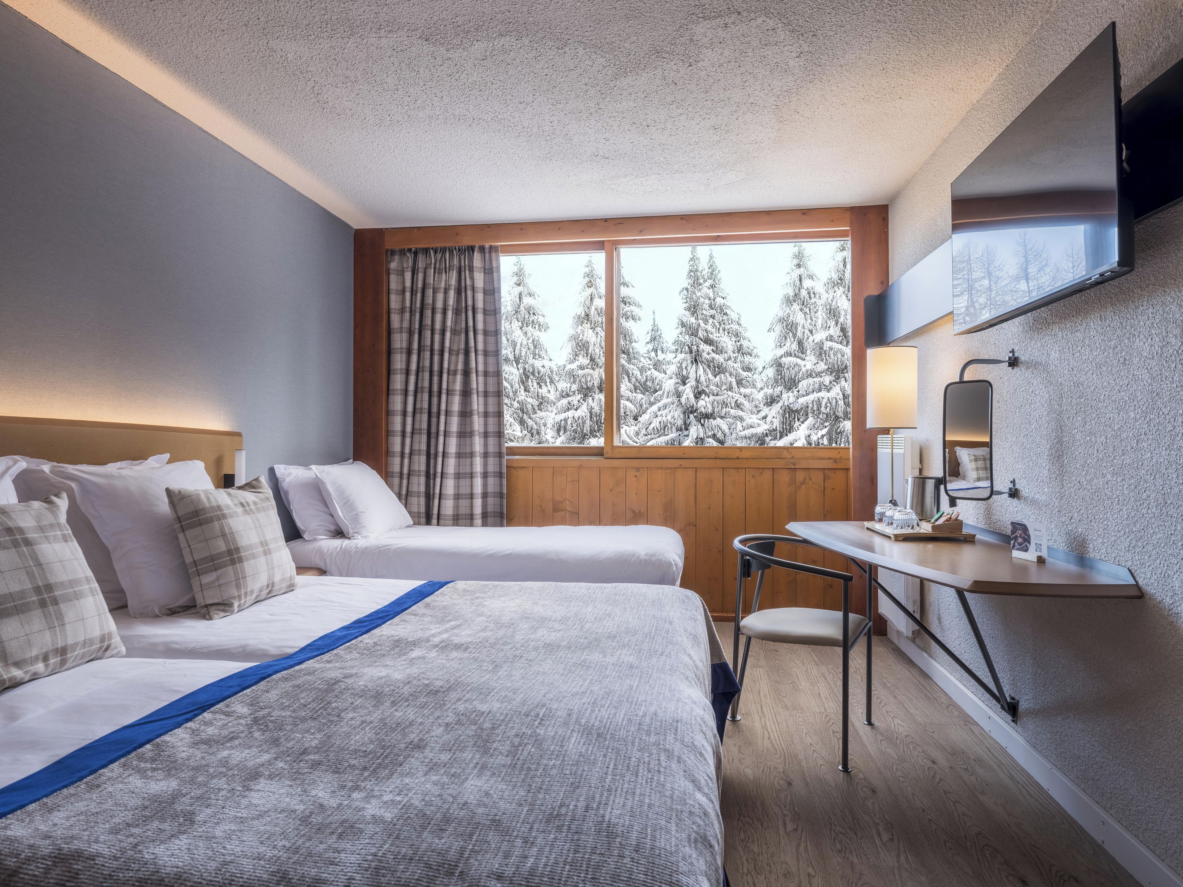A hotel room with two beds, a desk, a chair, a window with a view of snow-covered trees, and a mounted TV.