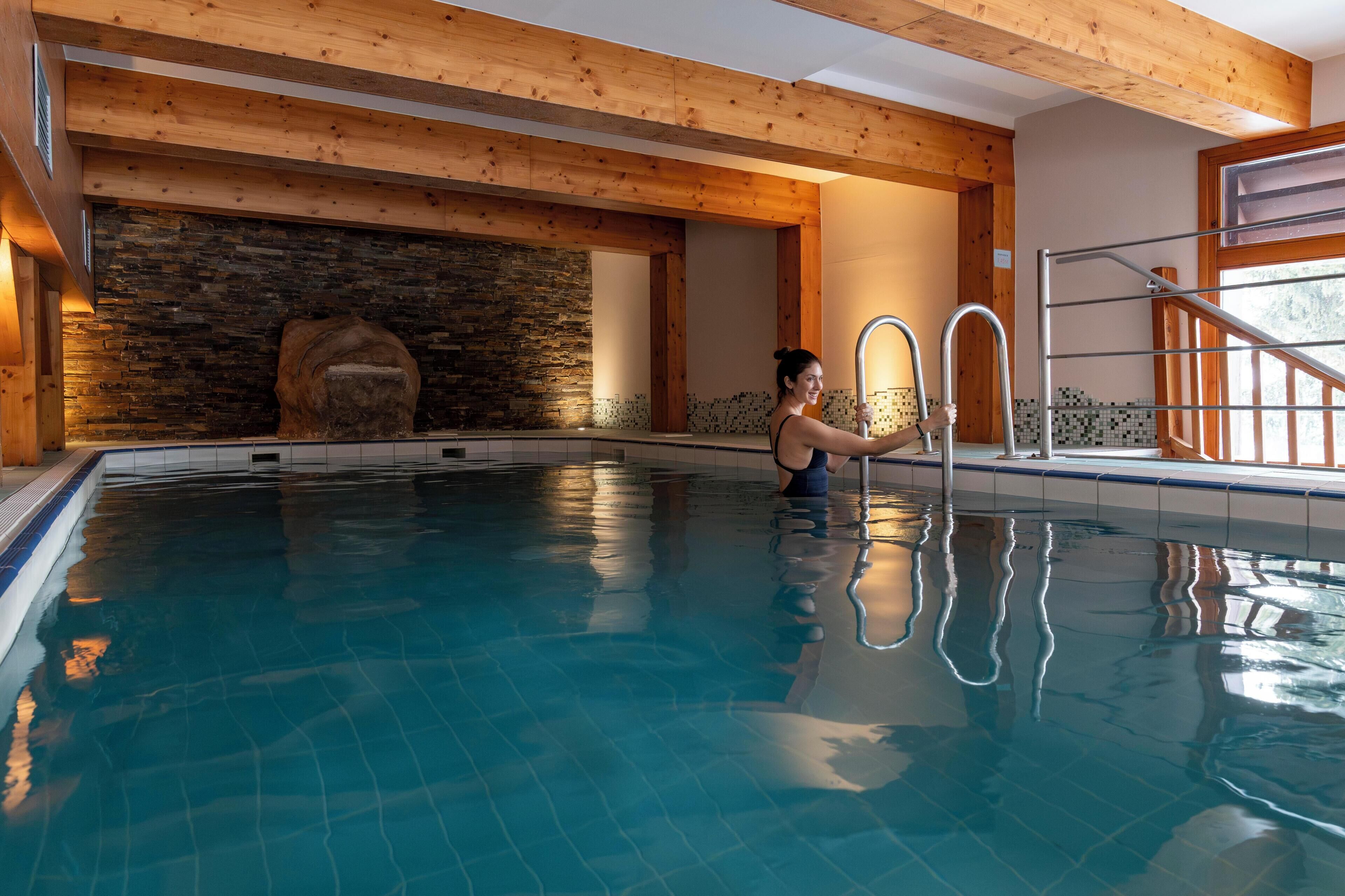 An indoor swimming pool with wooden beams, a stone wall, and a metal ladder.