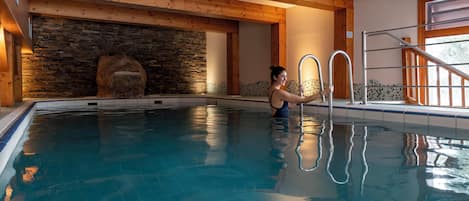 An indoor swimming pool with wooden beams, a stone wall, and a metal ladder.