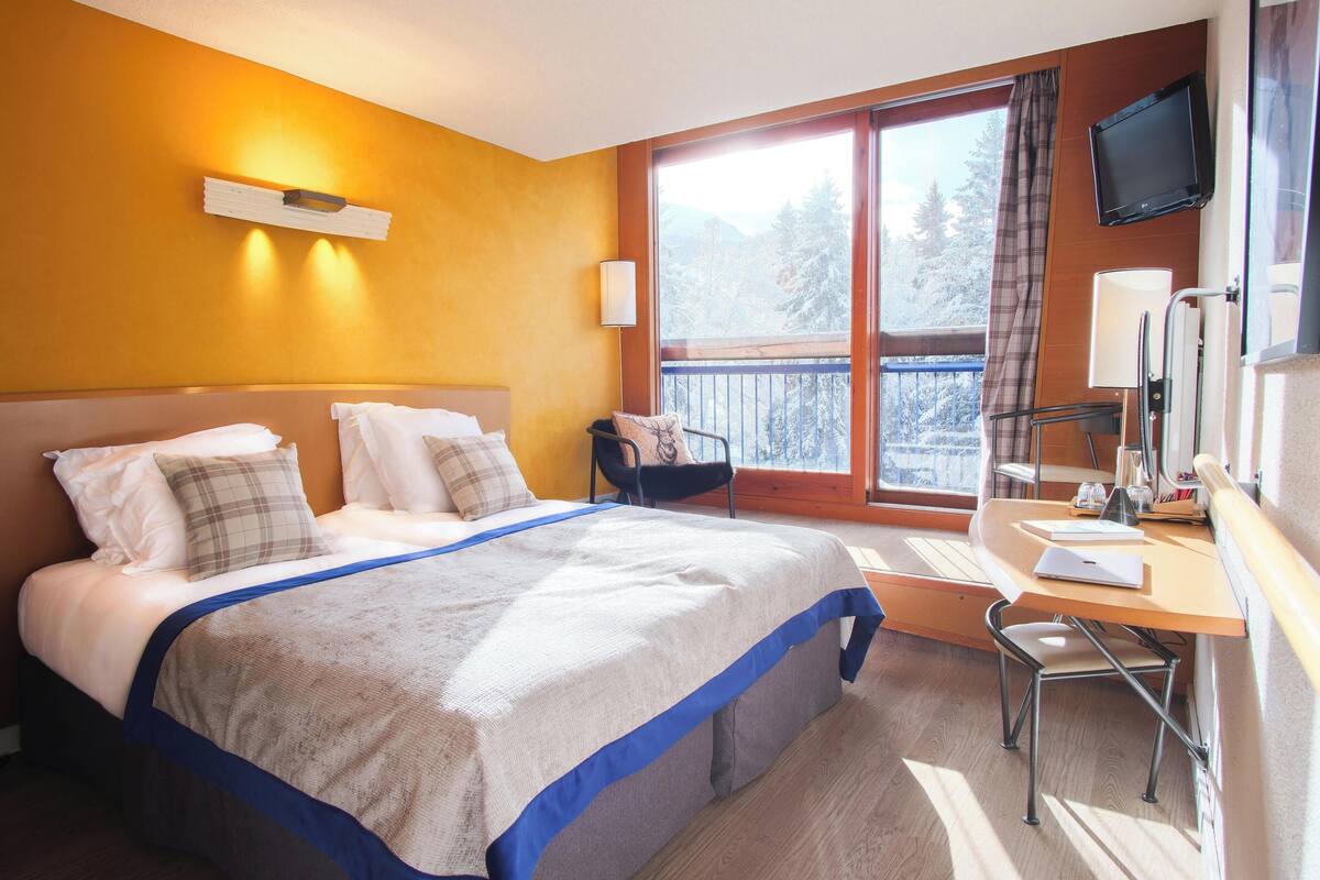 A hotel room with a bed, a desk, a chair, a TV, and a window with a view of trees.
