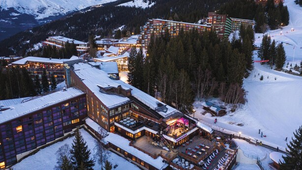 A snow-covered resort with multiple buildings, including a large lodge with a lit-up bar area and a ski slope in the background. - Belambra Clubs Arc 1800 - Hôtel Du Golf  (Bourg-Saint-Maurice)