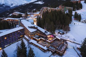 A snow-covered resort with multiple buildings, including a large lodge with a lit-up bar area and a ski slope in the background. - Belambra Clubs Arc 1800 - Hôtel Du Golf  (Bourg-Saint-Maurice)