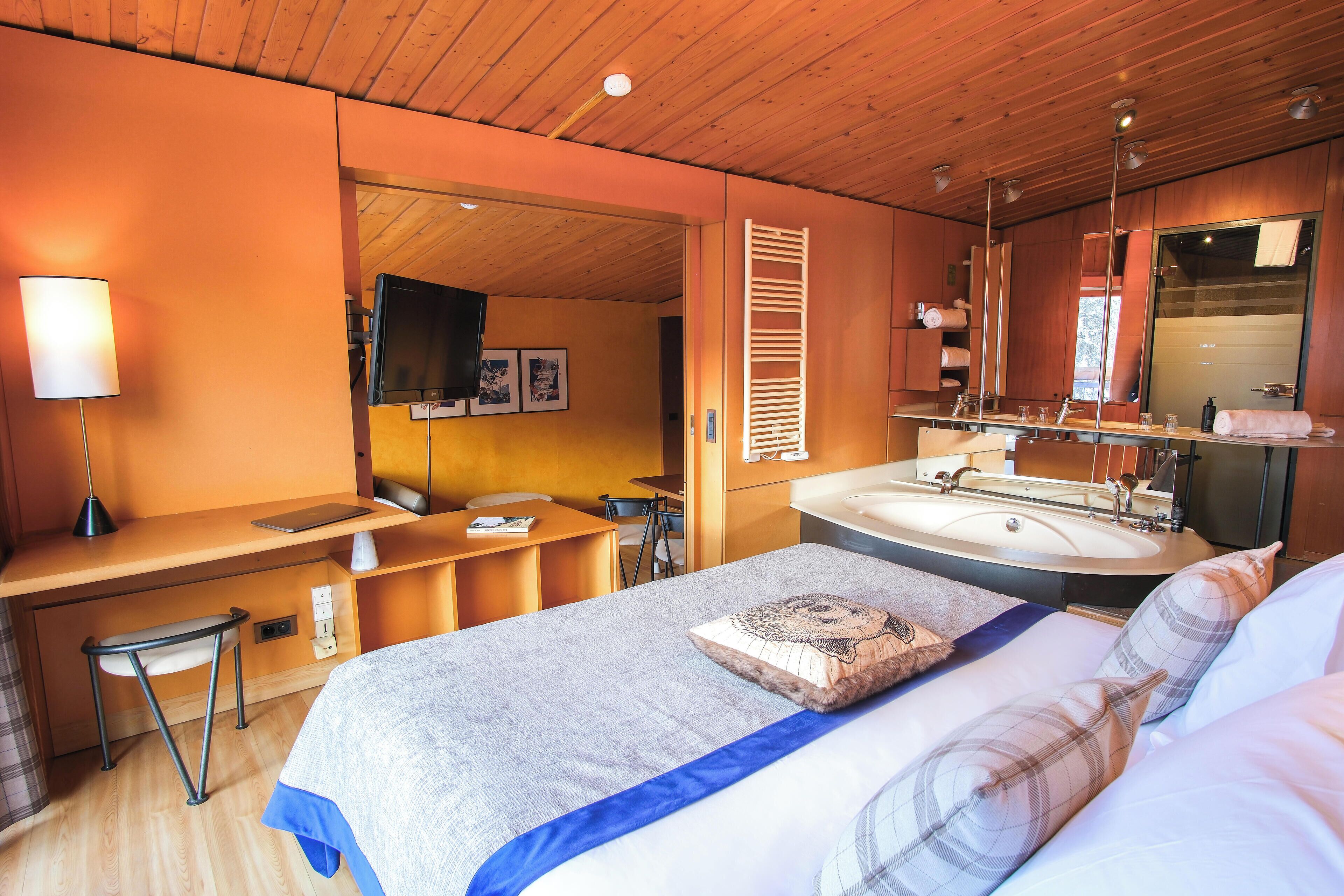 Habitación de hotel con techo de madera, cama con cubrecama azul, escritorio con silla, espejo grande y baño con lavamanos y espejo.