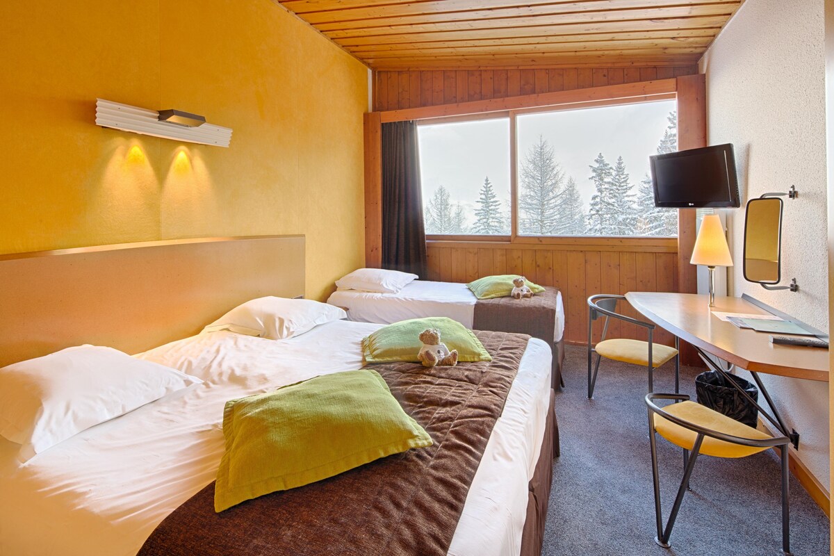 Habitación de hotel con dos camas, un escritorio, una silla, un televisor y una ventana con vista a árboles.