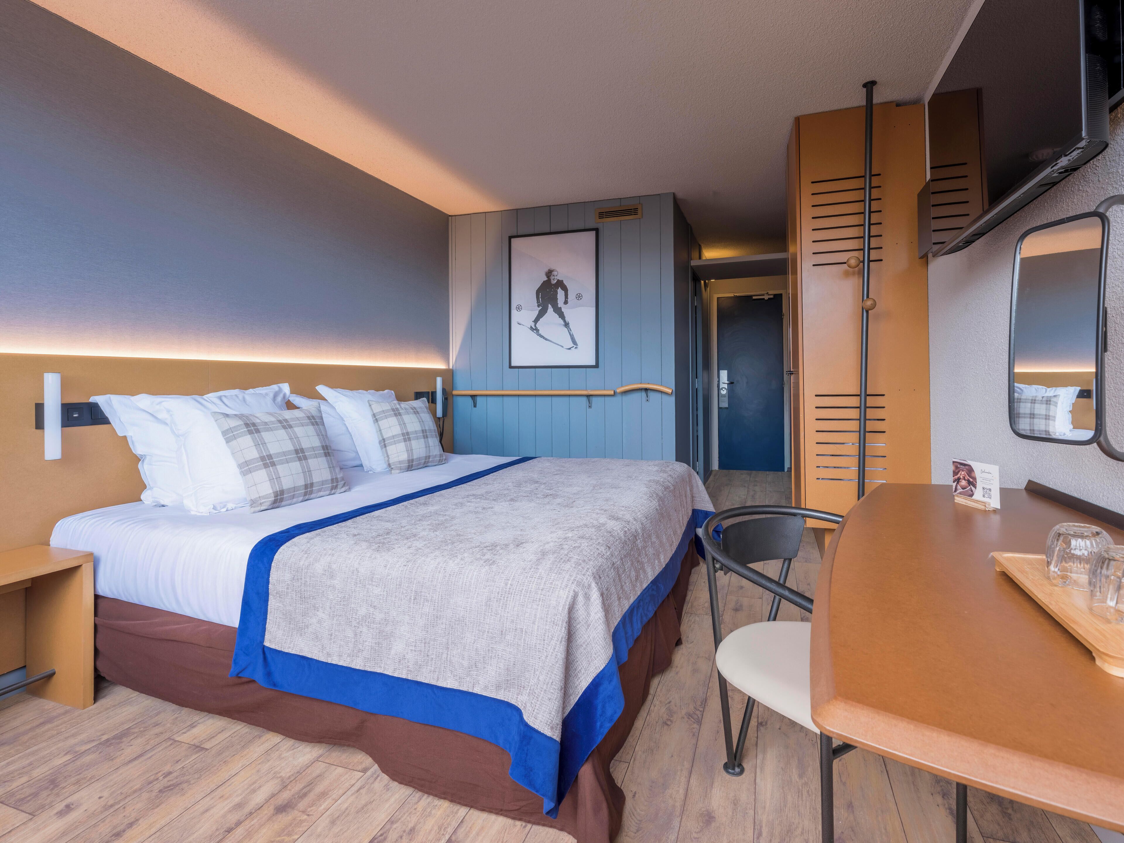 Camera d'albergo con un letto, una scrivania, una sedia, uno specchio, un quadro appeso al muro e una porta che dà su un'altra stanza.