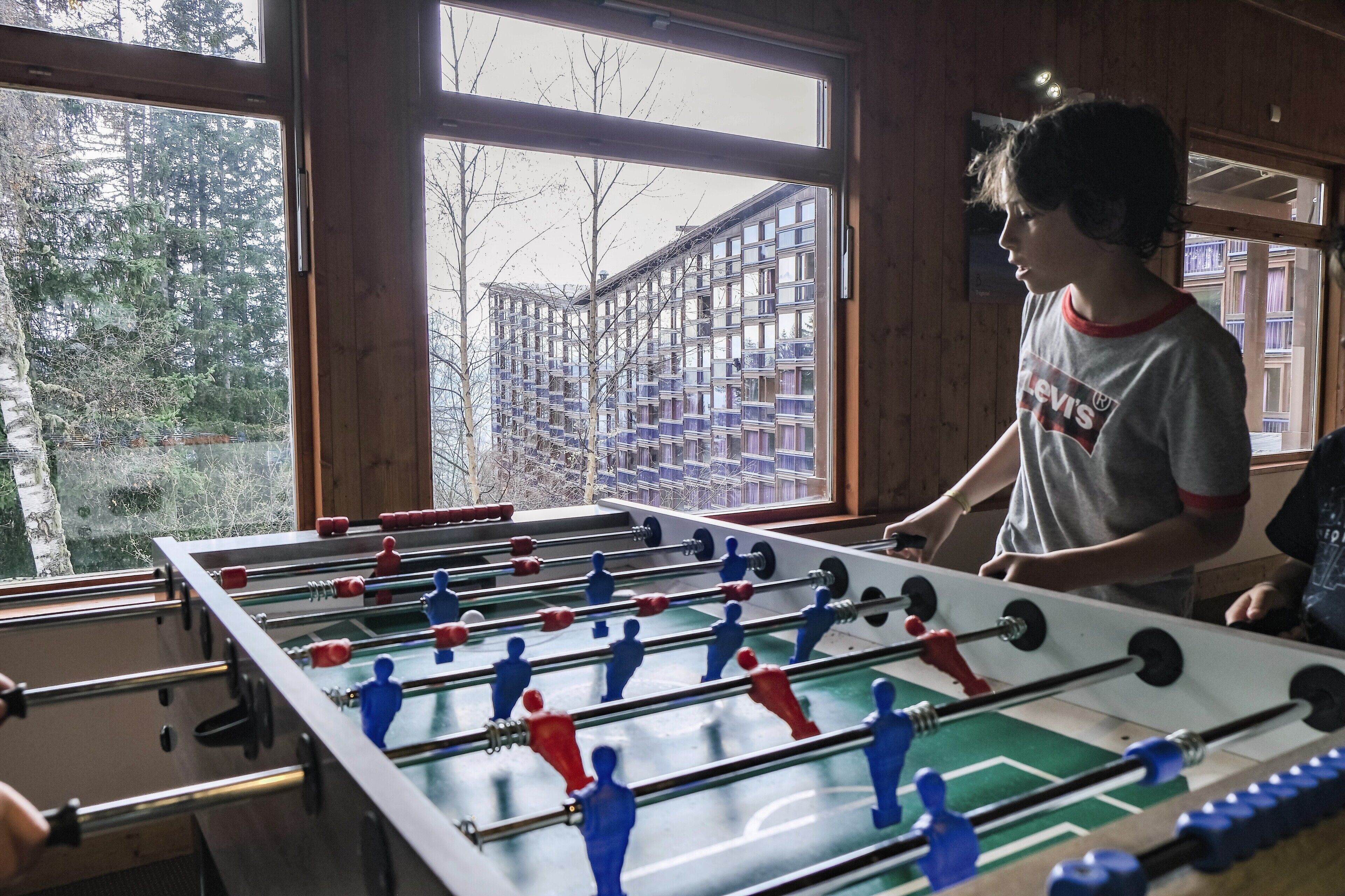 A foosball table in a room with large windows, wooden walls, and a view of a residential building.