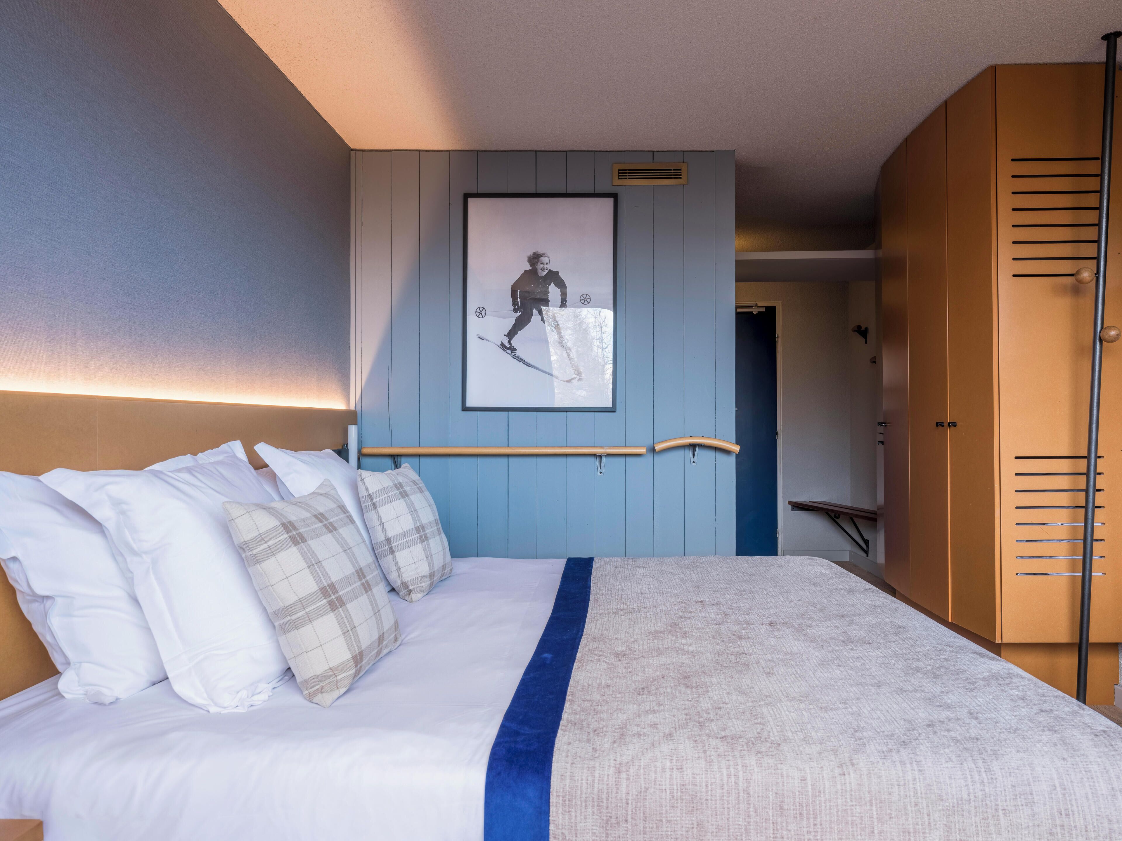 A neatly made bed with white and plaid pillows, a blue accent, and a framed picture of a skier on the wall.