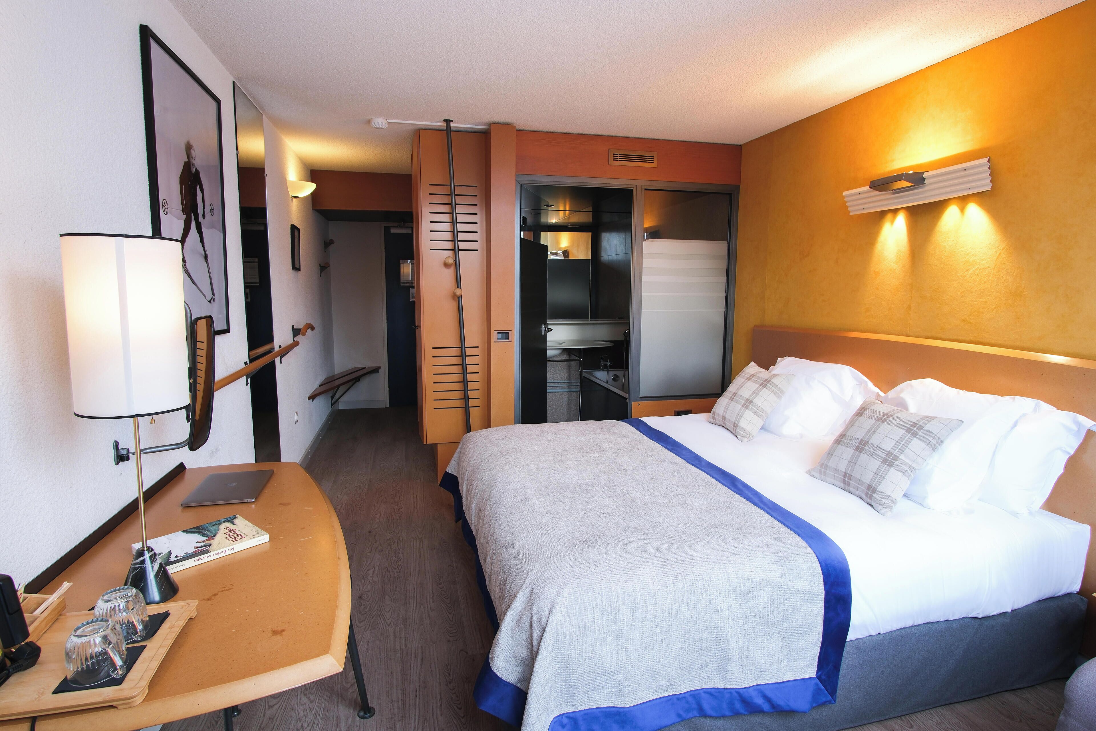 A hotel room with a bed, bedside table, lamp, and a door leading to a bathroom.