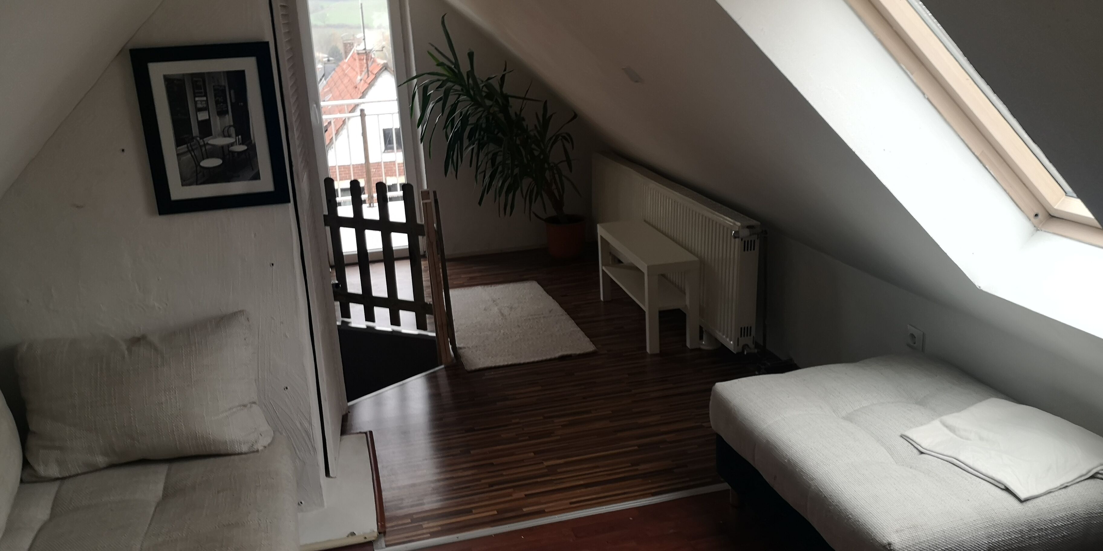 Ein kleiner, gemütlicher Raum mit schrägen Wänden, Holzboden, einem Bett, einem Stuhl, einem Heizkörper und einem Fenster.