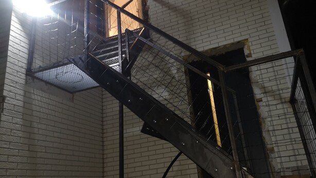 Una escalera metálica con barandilla de metal que conduce a un nivel superior de un edificio con paredes de ladrillo blanco.