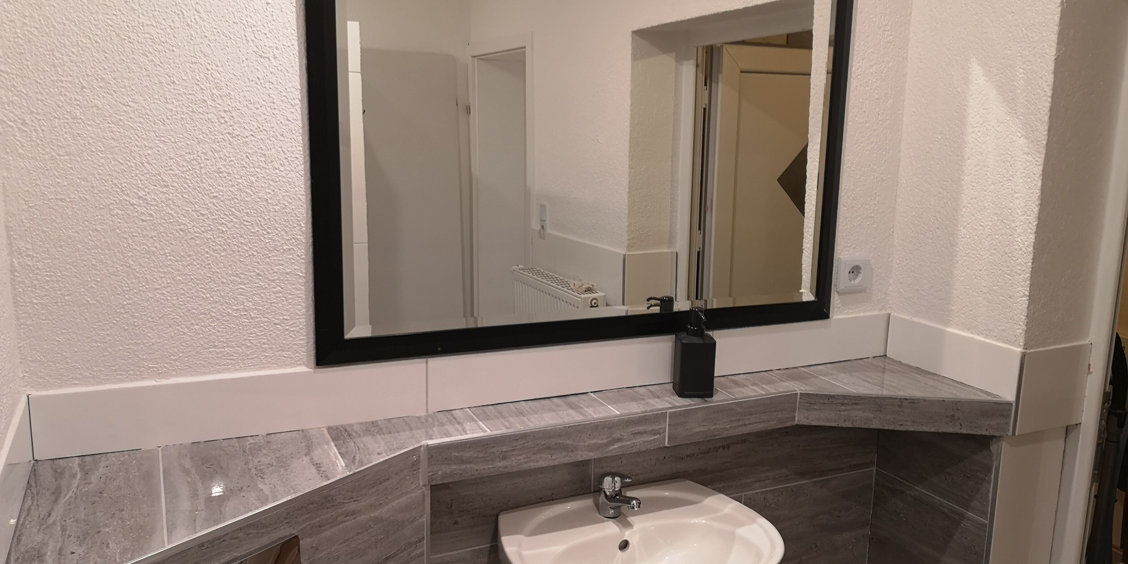 Un bany amb un lavabo blanc, un mirall gran i un dispensador de sabó negre.