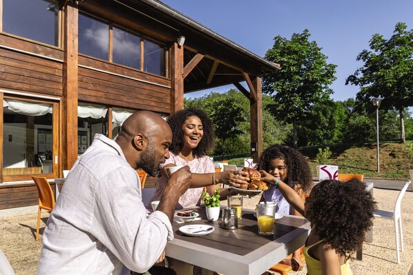 A family dining outdoors at a wooden building with large windows and a covered patio area. - Belambra Clubs Résidence Rocamadour - Les Portes De Dordogne (Alvignac)
