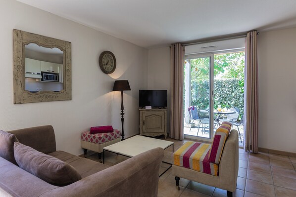 A living room with a sofa, a small table, a television, and a view of an outdoor patio with a table and chairs. - Belambra Clubs Résidence Rocamadour - Les Portes De Dordogne (Alvignac)