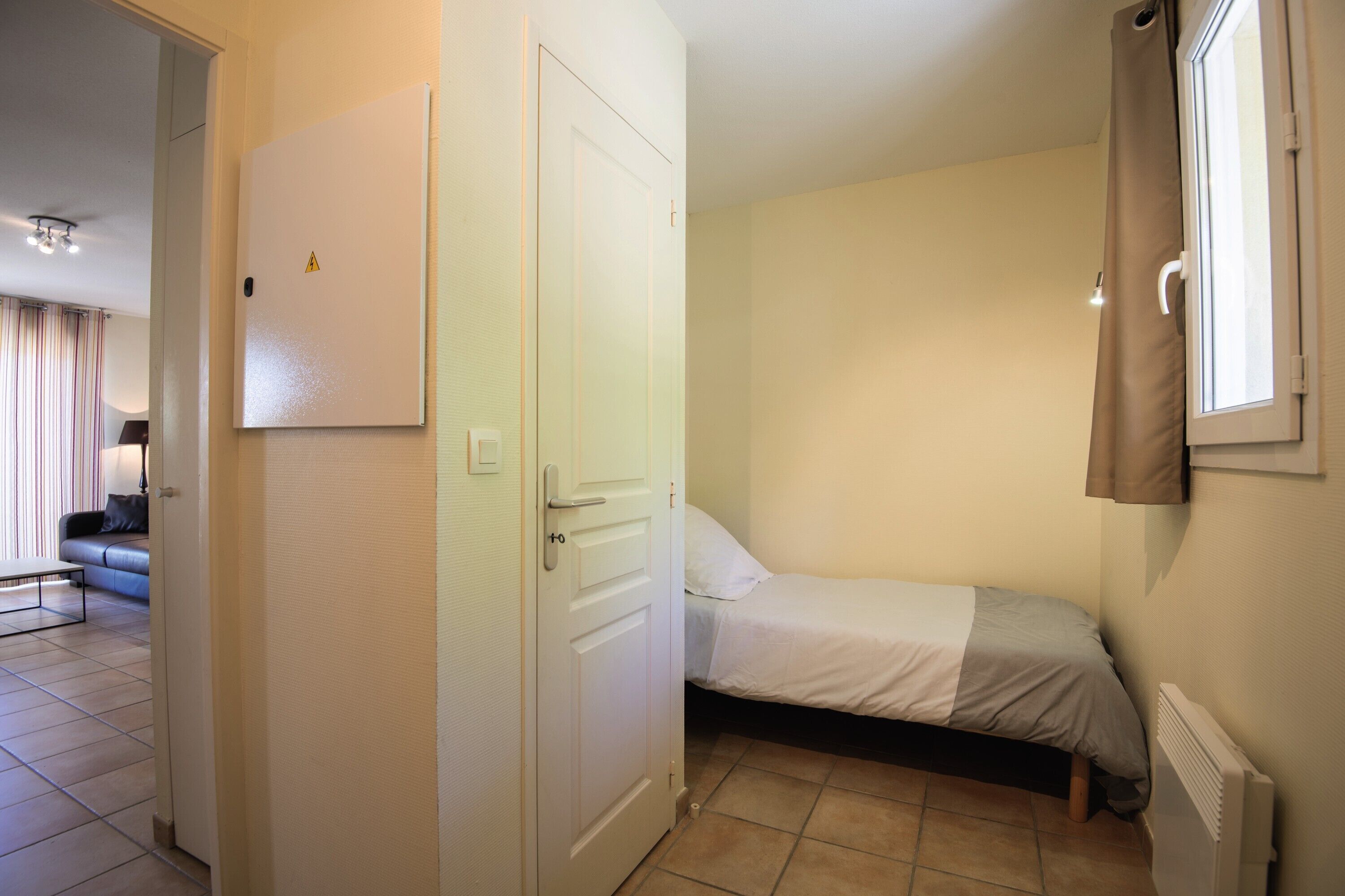 Un dormitorio pequeño y bien iluminado con una cama individual, un radiador y una puerta que da a un salón con un sofá y una mesa.