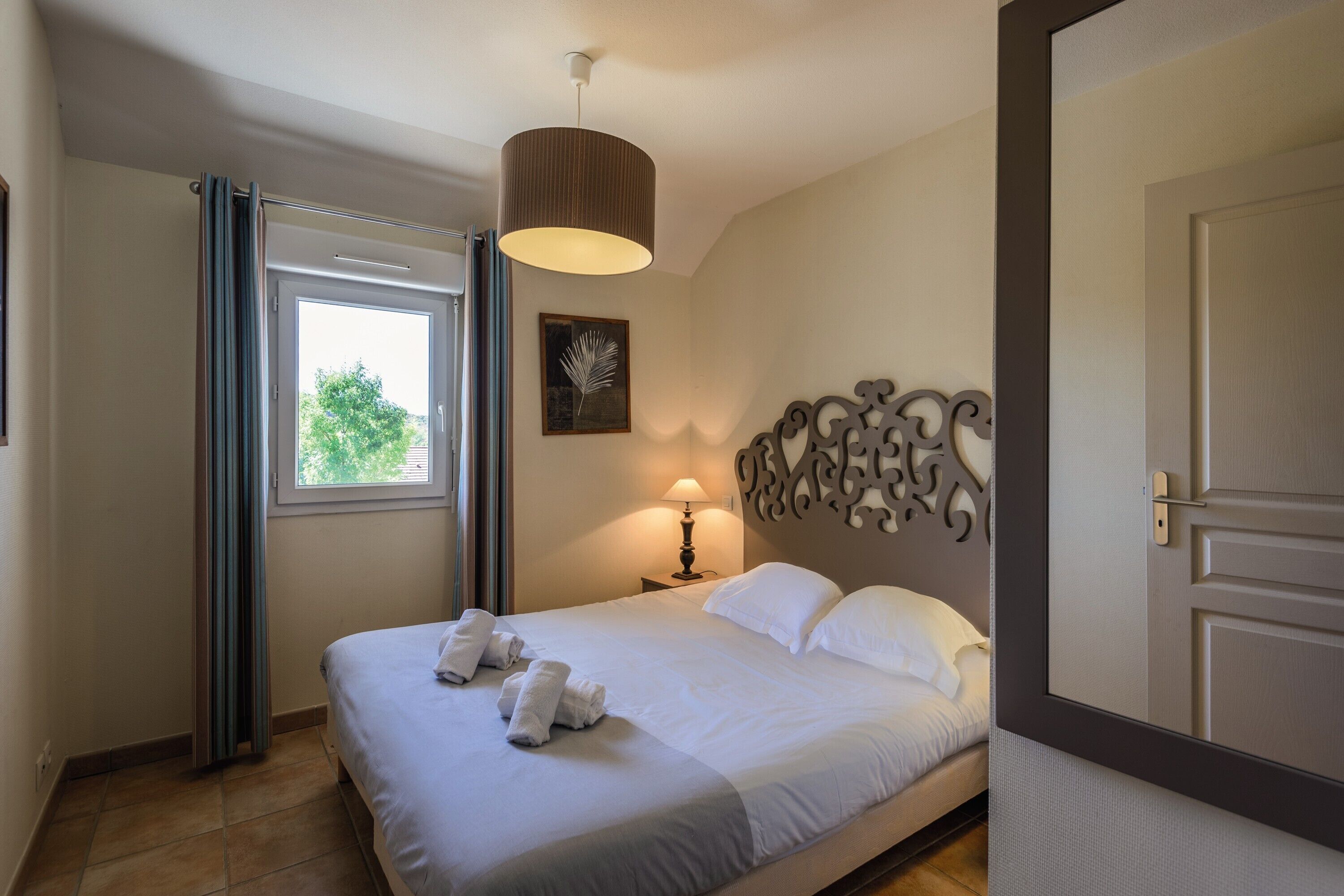 Una cama bien hecha con sábanas y toallas blancas, una lámpara de noche, un adorno en la pared y una ventana con cortinas.