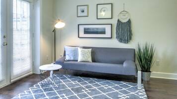 A living room with a gray sofa, a small round table, and a patterned rug. There are two framed pictures and a dreamcatcher on the wall.