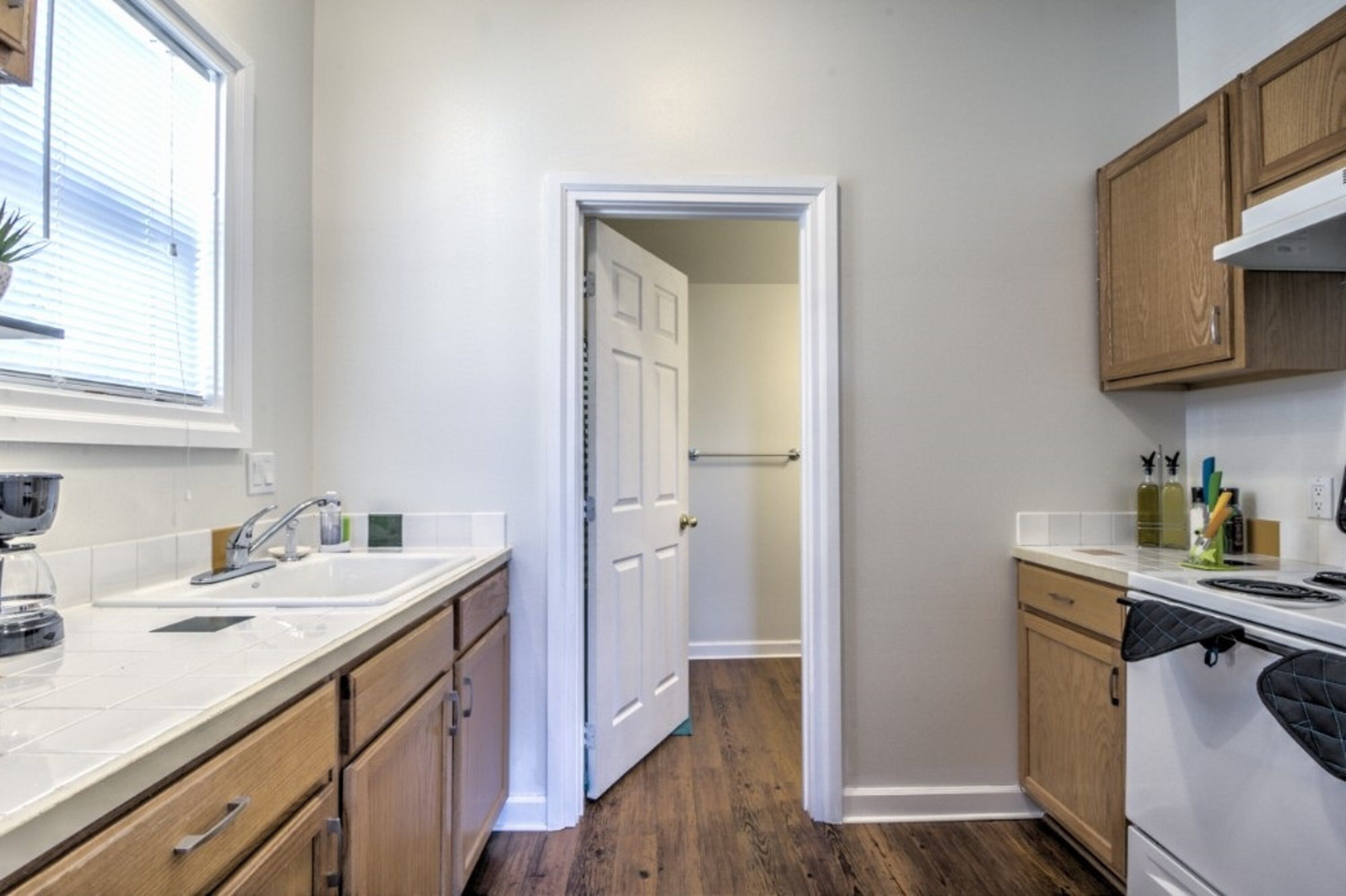 A narrow kitchen with wooden cabinets, a white countertop, and a window with blinds.