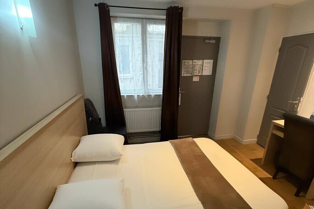 A hotel room with a bed, a desk, a chair, a window with curtains, and a door with a bulletin board.