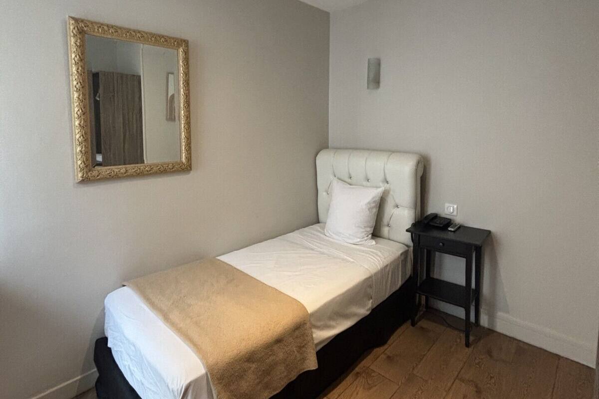 A bedroom with a single bed, a bedside table, a nightstand, a mirror, and a light fixture.