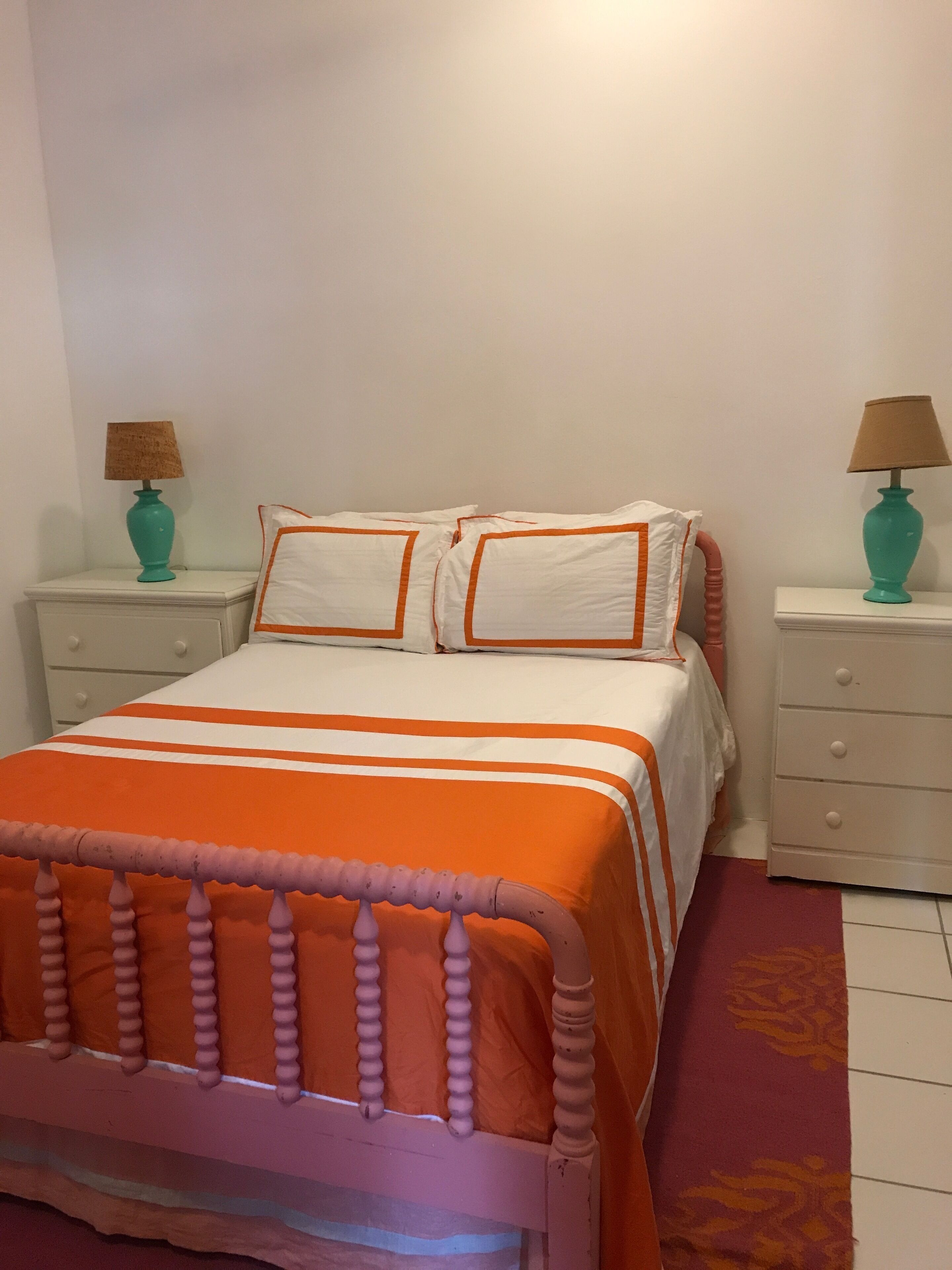 Una cama con una colcha naranja y blanca a rayas, dos almohadas blancas con ribete naranja, y un cabecero de madera. Hay dos mesitas de noche con lámparas azul verdosas y un cajón blanco.
