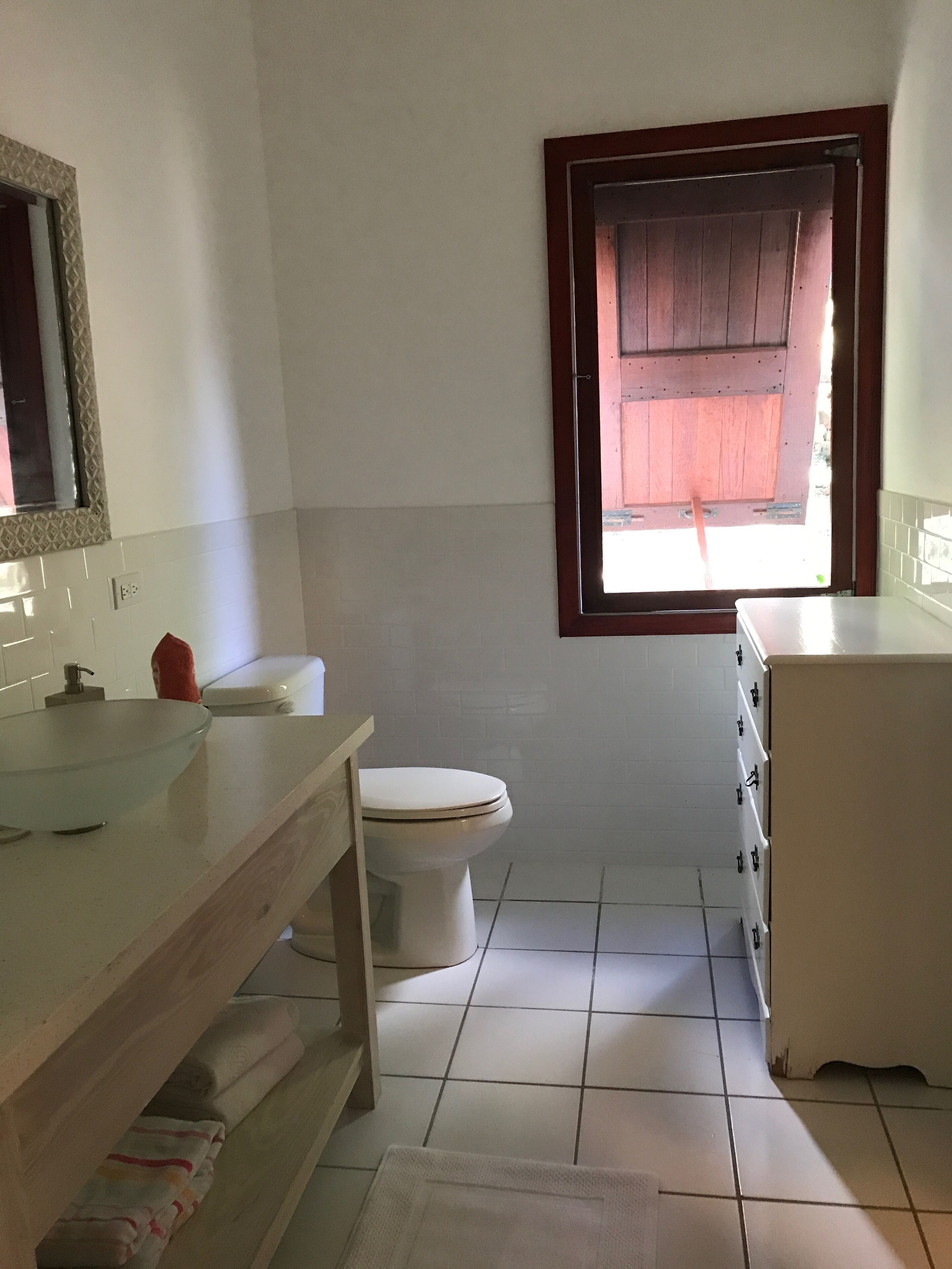 Een badkamer met een glazen wastafel, toilet, houten wastafelmeubel en een raam met houten kader.