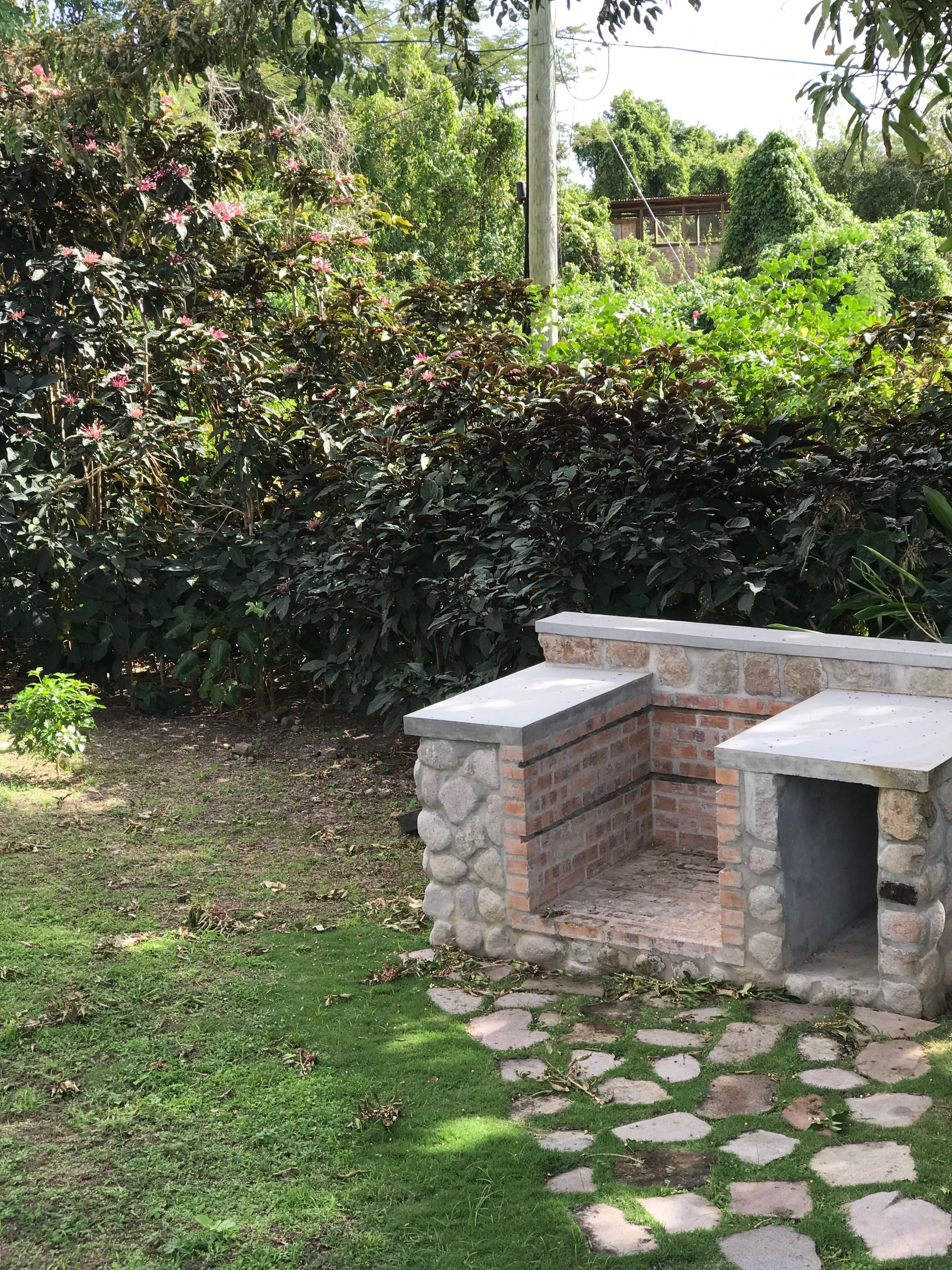 A stone barbecue area with a brick chimney, surrounded by a garden with various plants and a stone pathway.