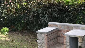 A stone barbecue area with a brick chimney, surrounded by a garden with various plants and a stone pathway.
